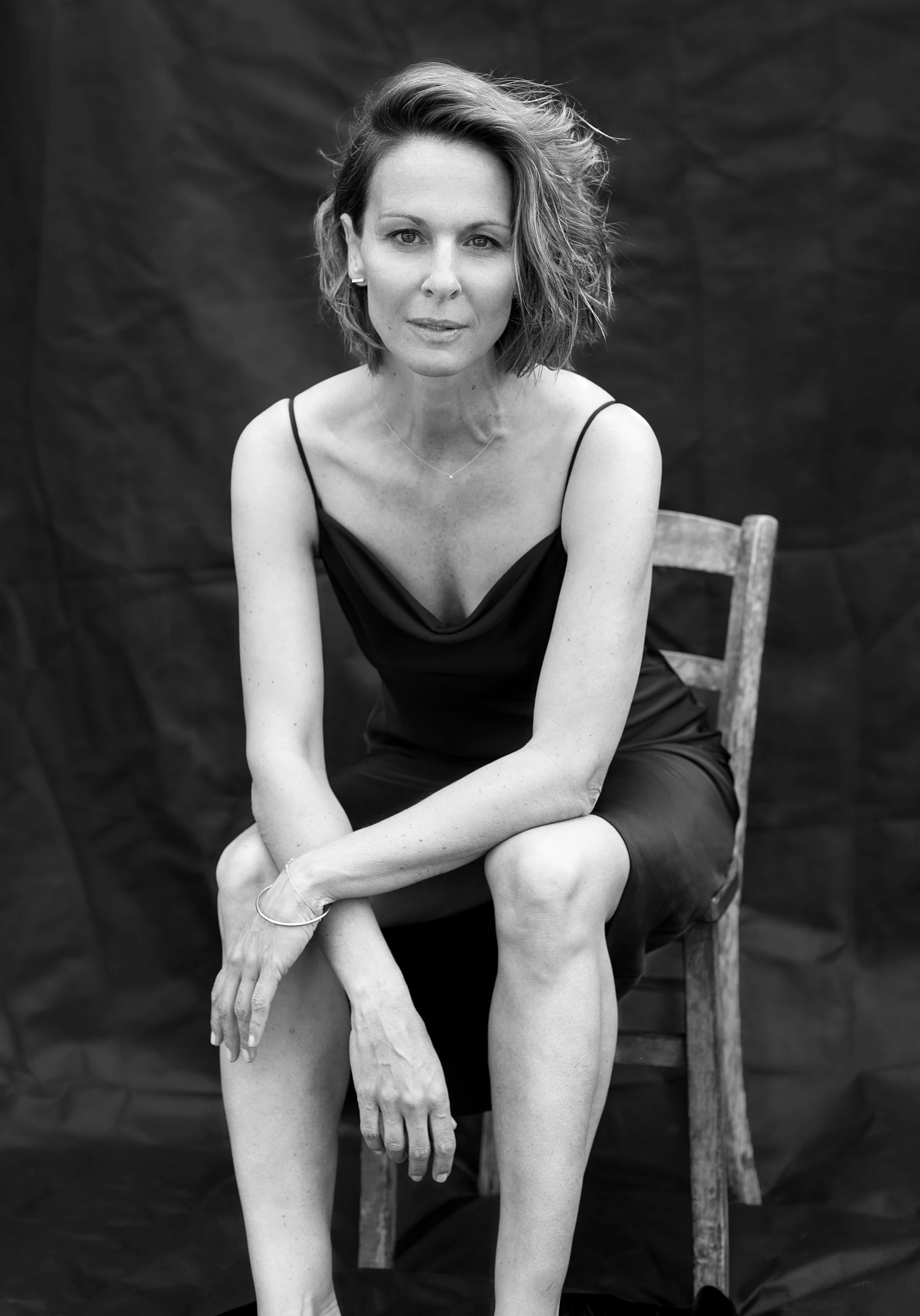Schwarz-Weiß-Fotografie einer Frau in einem schwarzen Kleid, sitzend auf einem Holzstuhl, vor einem dunklen Hintergrund, mit kurzen, leicht gelockten Haaren und einem neutralen Gesichtsausdruck.