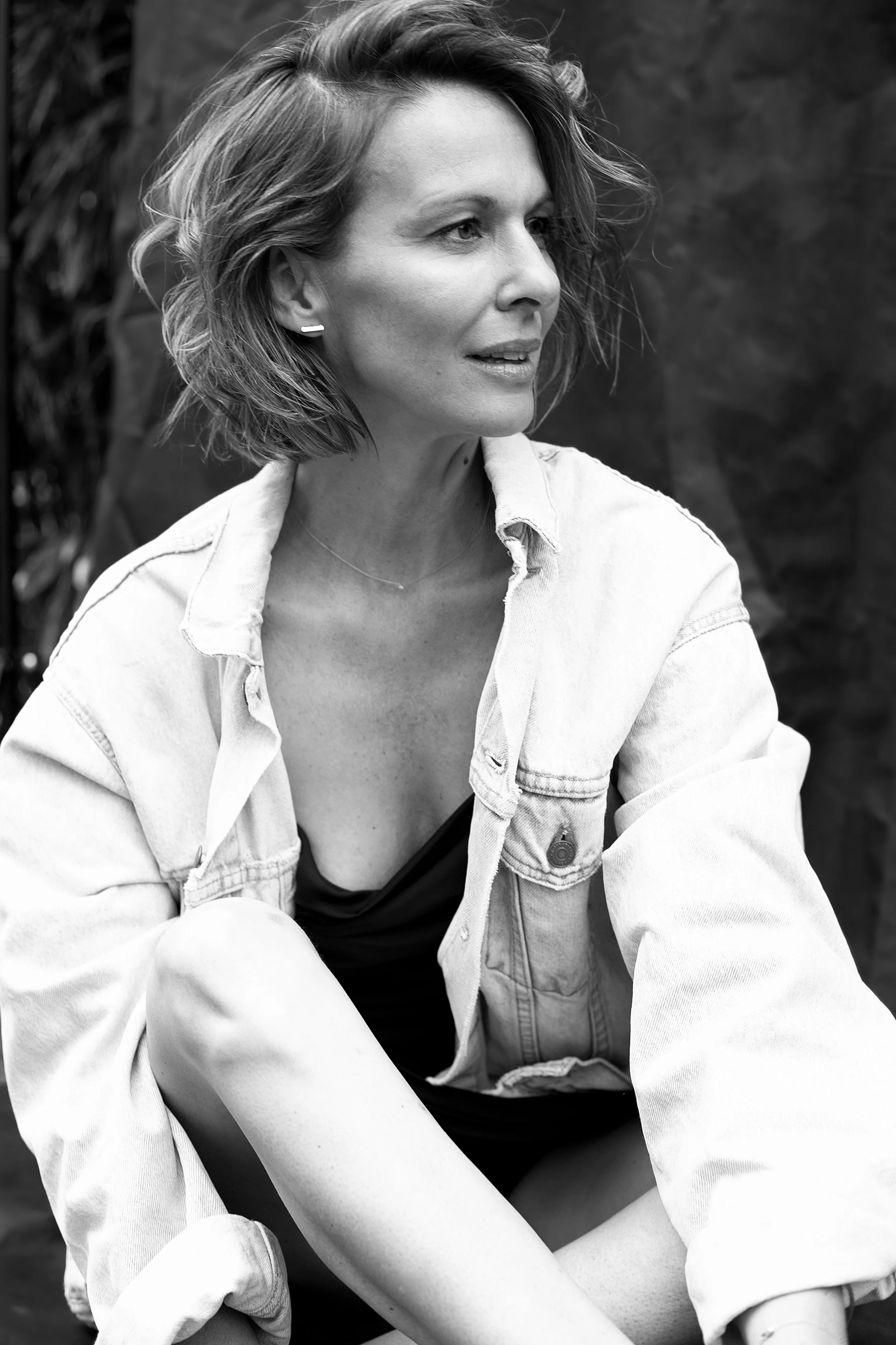 Schwarz-weiß-Fotografie einer Frau mit lockigem, schulterlangem Haar, die in einer sitzenden Haltung nach rechts schaut, trägt eine Jeansjacke und ein dunkles Kleid, umgeben von Pflanzen im Hintergrund.