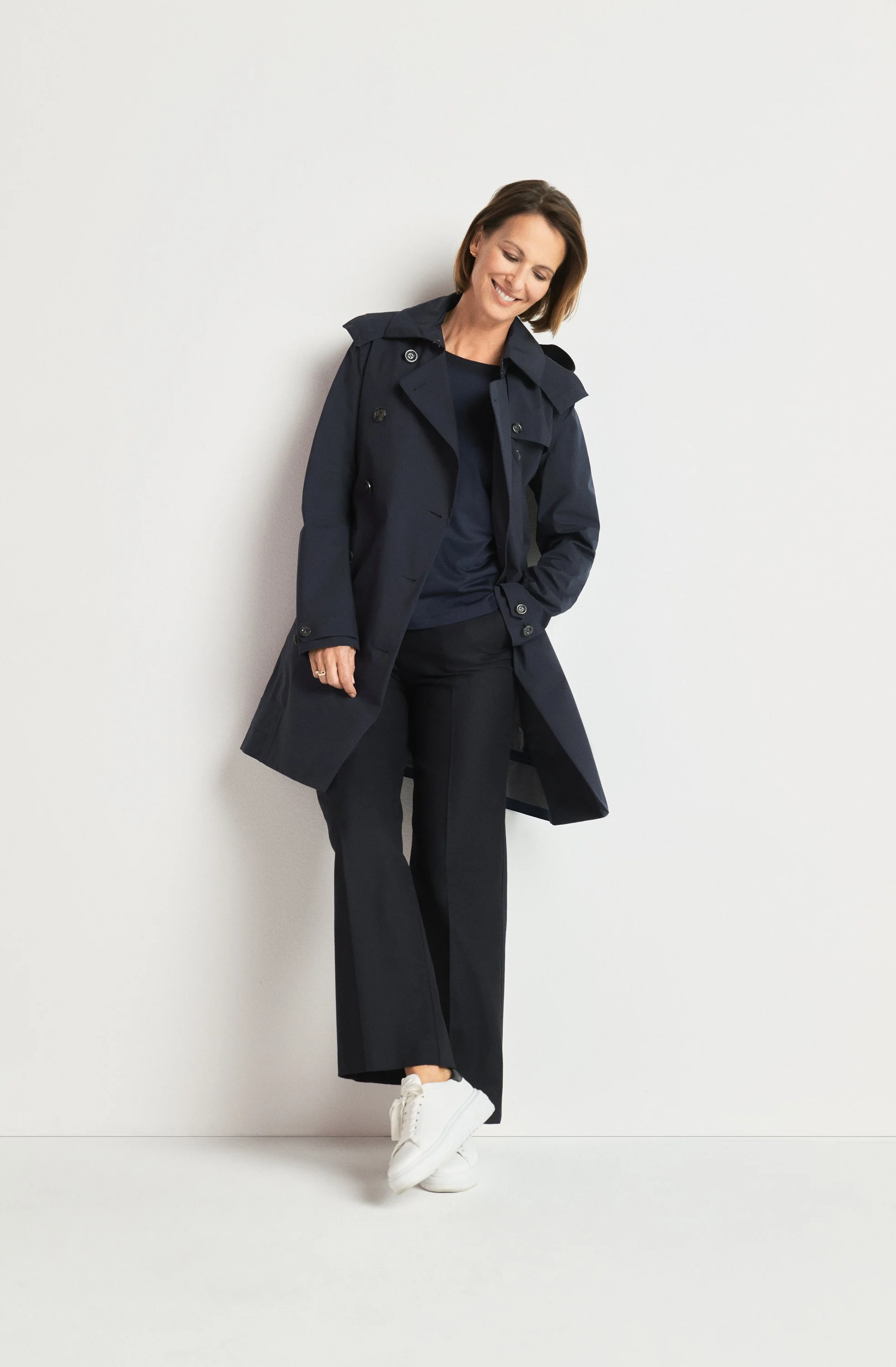Eine Frau trägt eine dunkelblaue Jacke, eine schwarze Hose und weiße Sneakers, und steht vor einer weißen Wand.