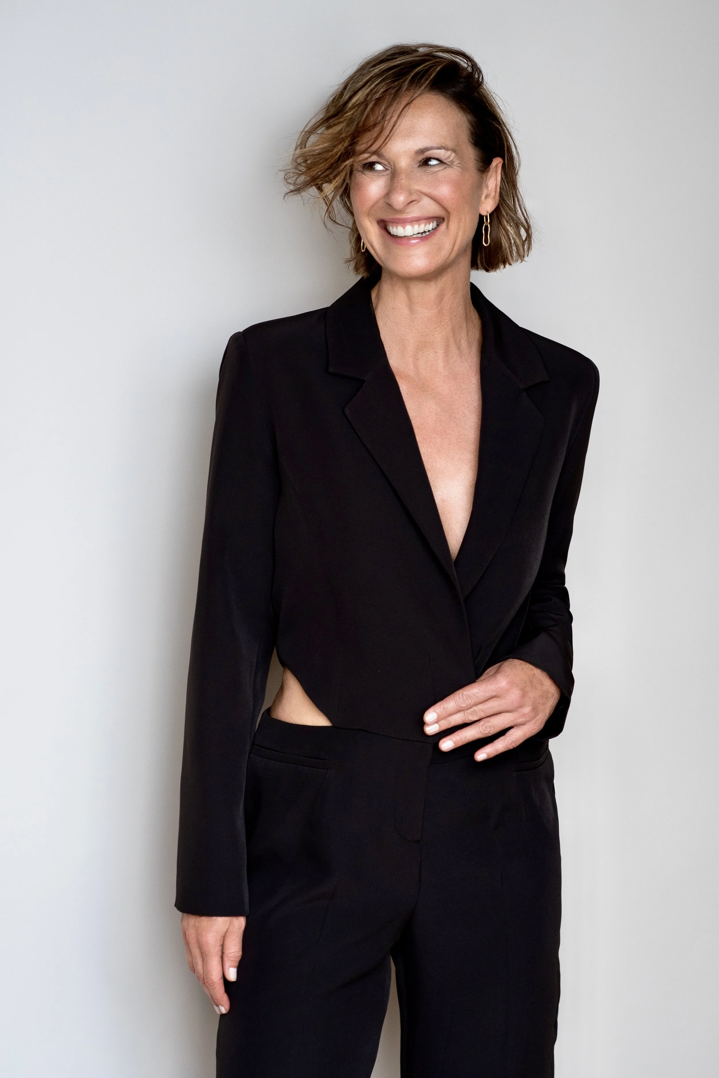 Eine lachende Frau trägt einen schwarzen Anzug, ihr Haar ist kurz und gewellt, vor hellem Hintergrund.