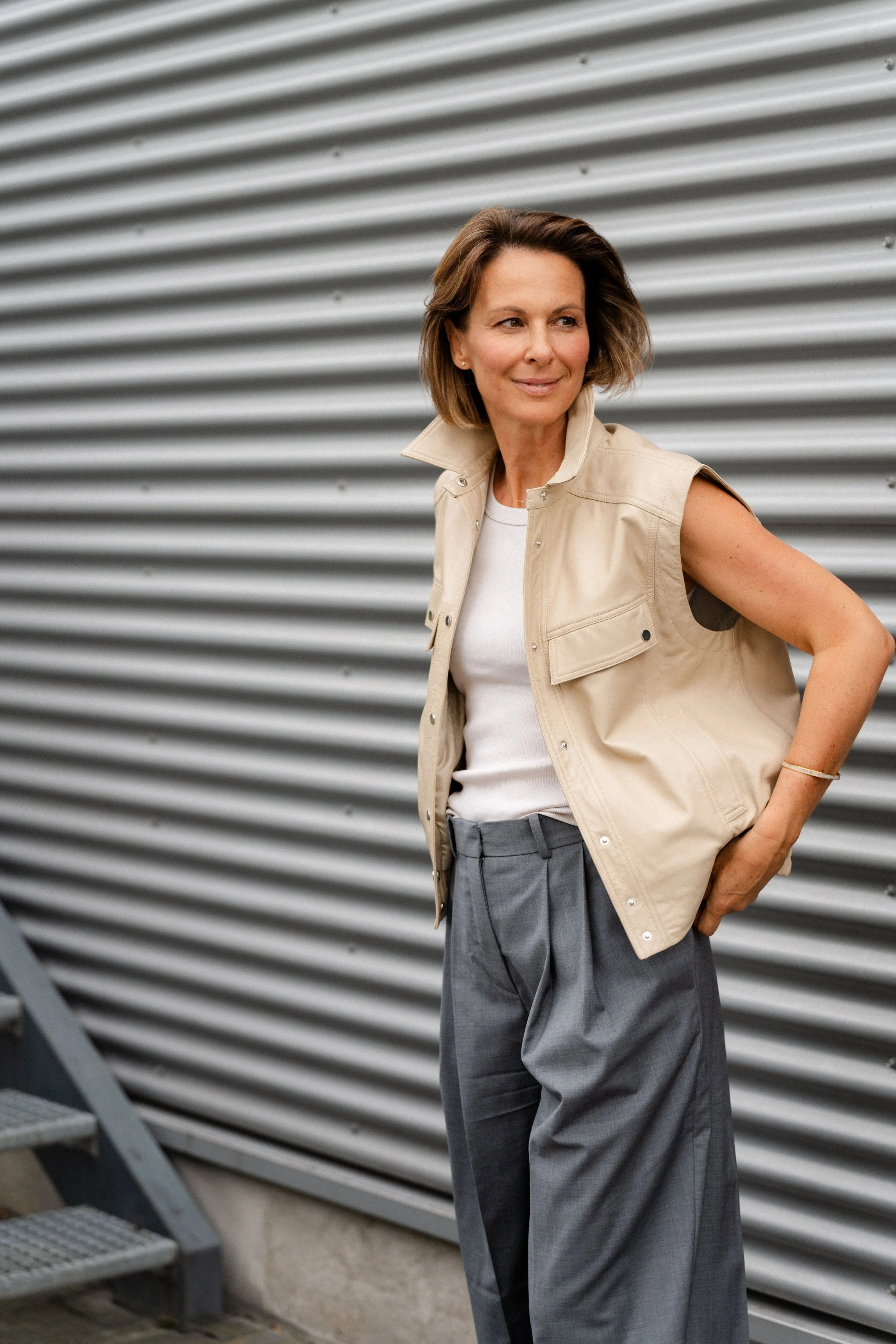 Frau steht vor einer metallverkleideten Wand, trägt eine beige Weste, weiße T-Shirt und graue Hosen, schaut seitlich, lächelt leicht.