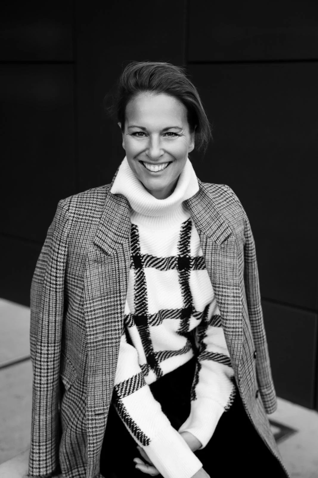 Schwarz-Weiß-Foto einer lächelnden Frau mit kurzen Haaren, die einen karierten Mantel trägt, in einem modernen Innenraum.