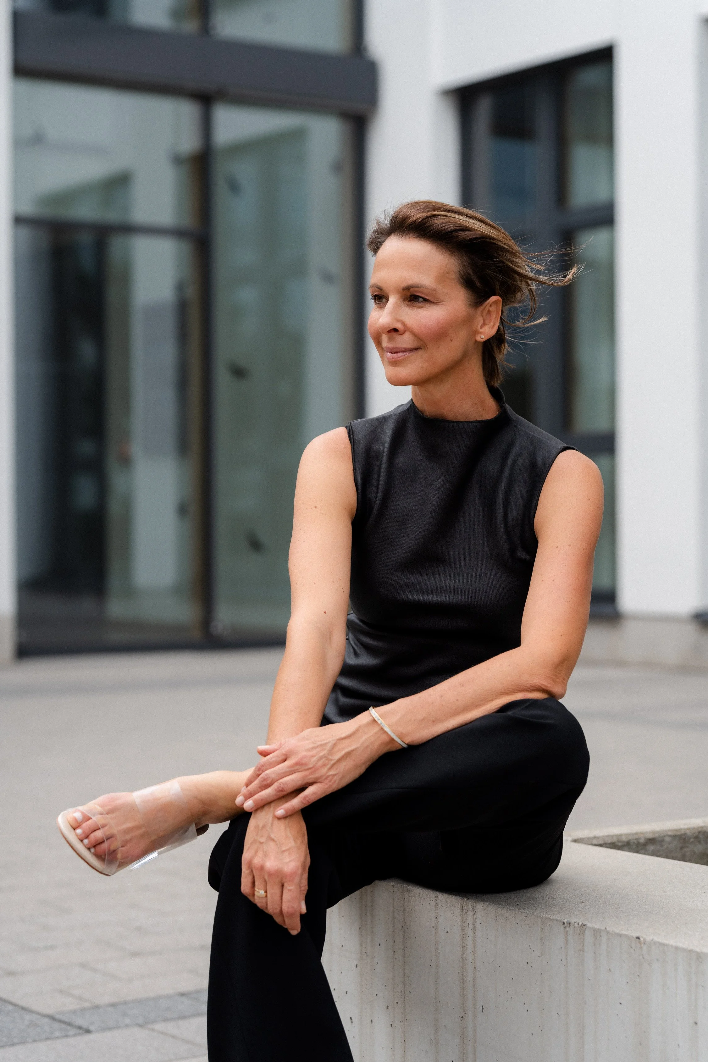 Eine Frau sitzt auf einer Betonbank vor einem modernen Gebäude, trägt ein schwarzes Kleid und offene Schuhe, schaut nach rechts, lächelt leicht und scheint entspannt zu sein.
