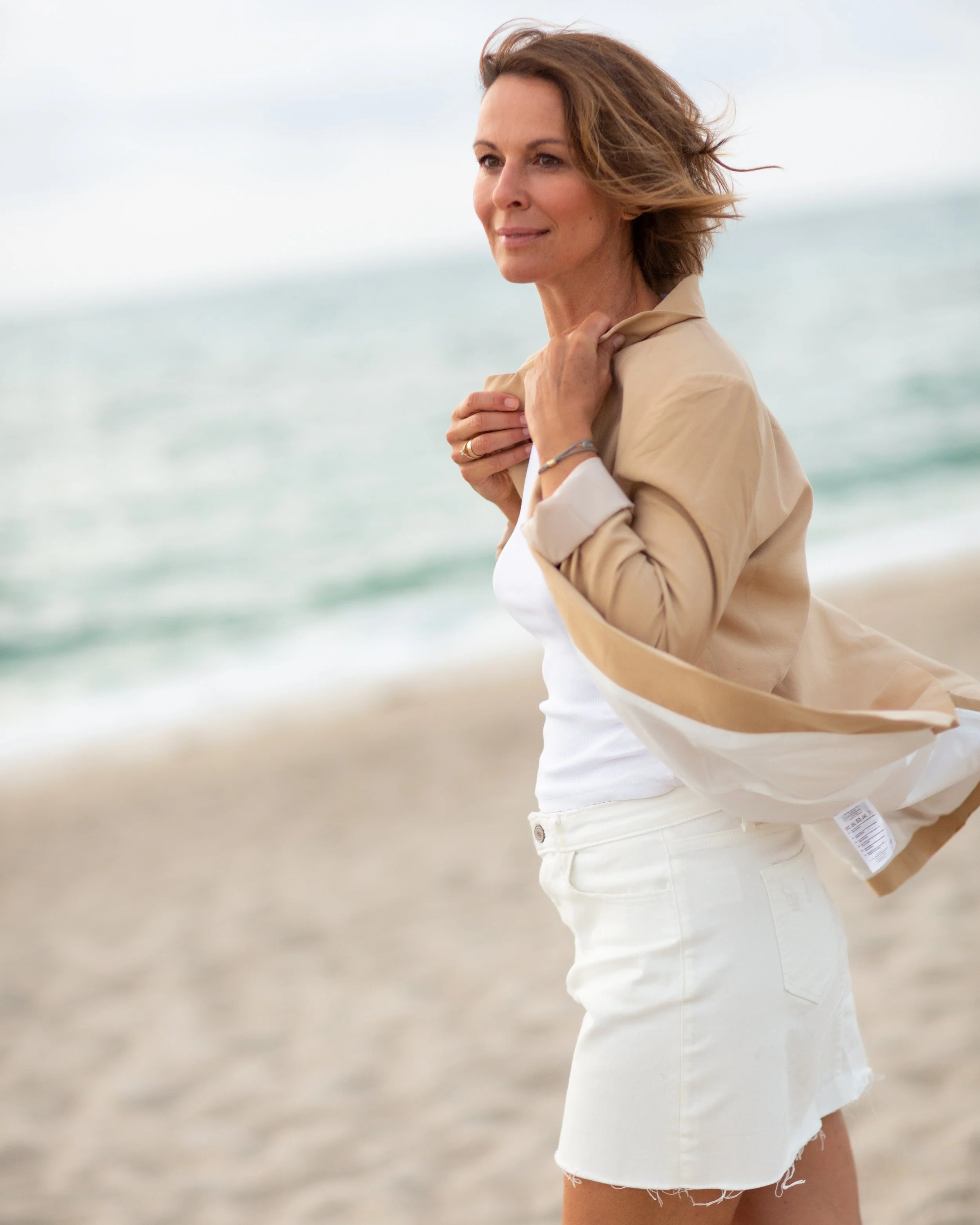 Eine Frau steht am Strand bei windigem Wetter, trägt eine beige Jacke, weiße Shorts und eine weiße Top, während sie an der Küste steht.