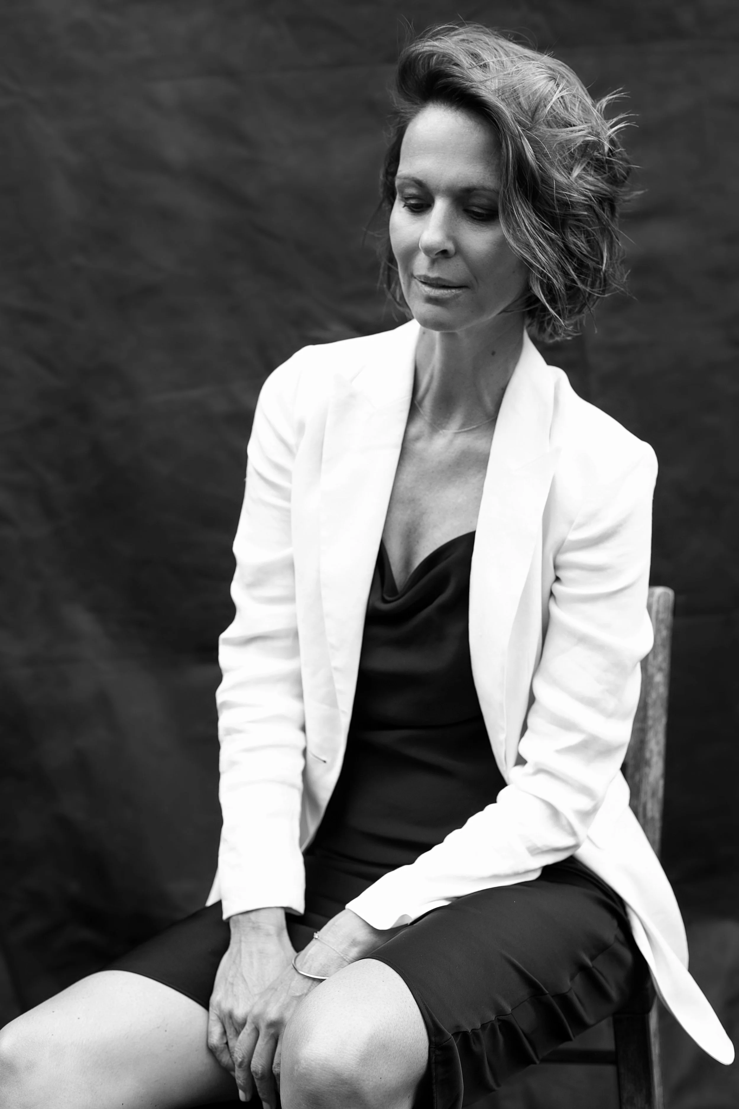 Schwarz-weiß Fotografie einer Frau mit kurzen, lockigen Haaren, die in einem weißen Blazer und schwarzem Kleid sitzt. Sie schaut nach unten mit nachdenklicher Miene vor einem dunklen, strukturierten Hintergrund.