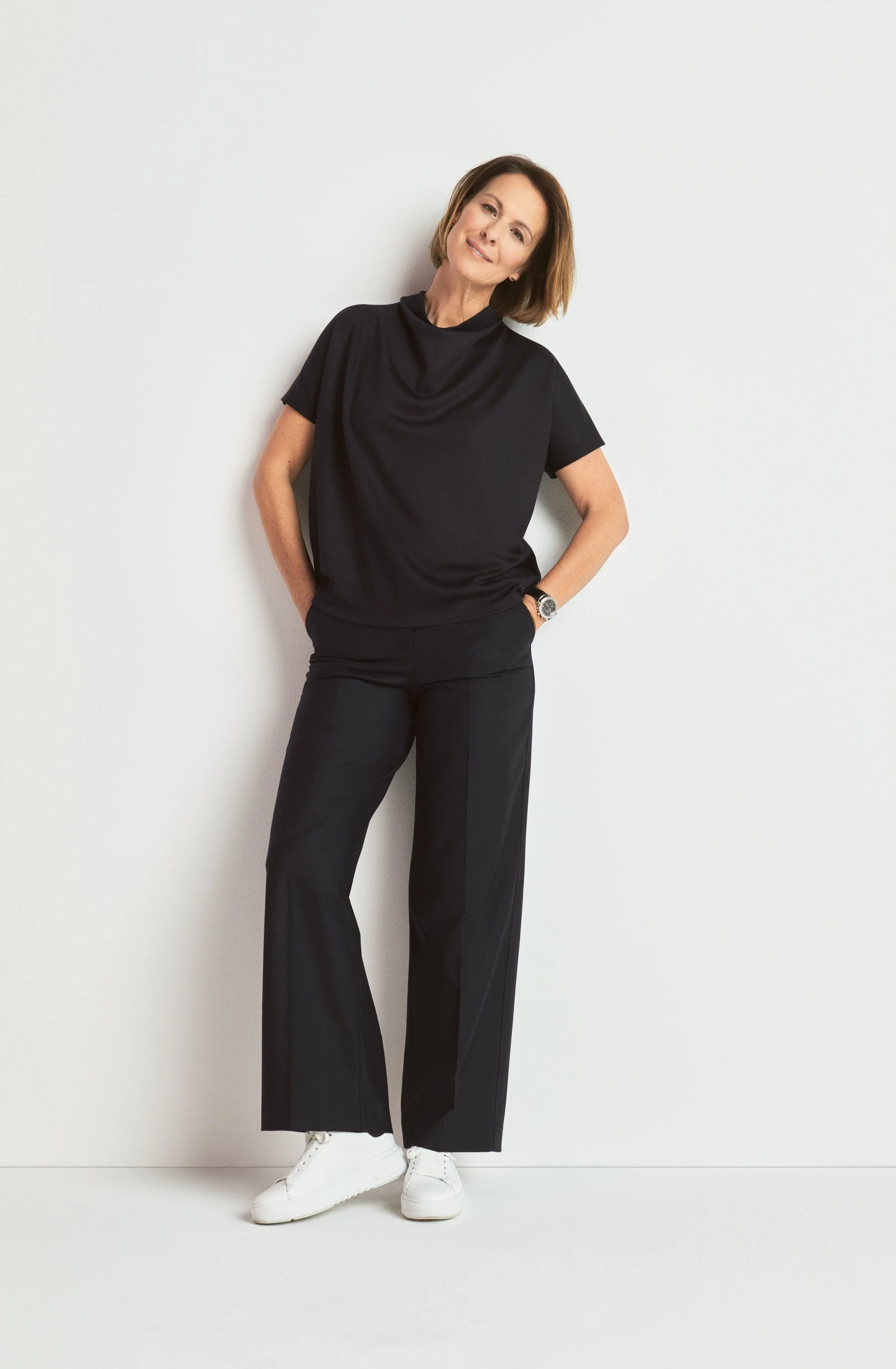 Frau in schwarzer Kleidung steht vor weißer Wand, lächelt und hat kurze braune Haare, trägt weißen Sneakers, Uhren am Handgelenk