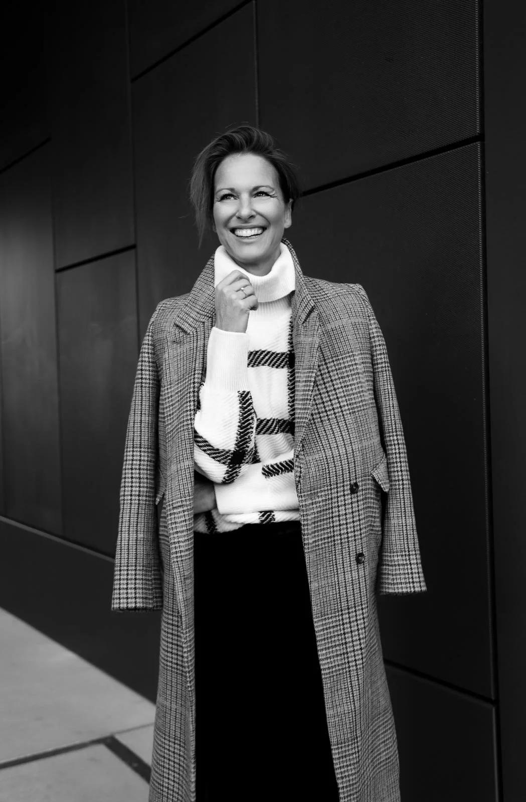 Eine lachende Frau mit kurzen Haaren trägt einen karierten Mantel über einem gestreiften Pullover und steht vor einer dunklen Wand in Schwarzweiß fotografiert.