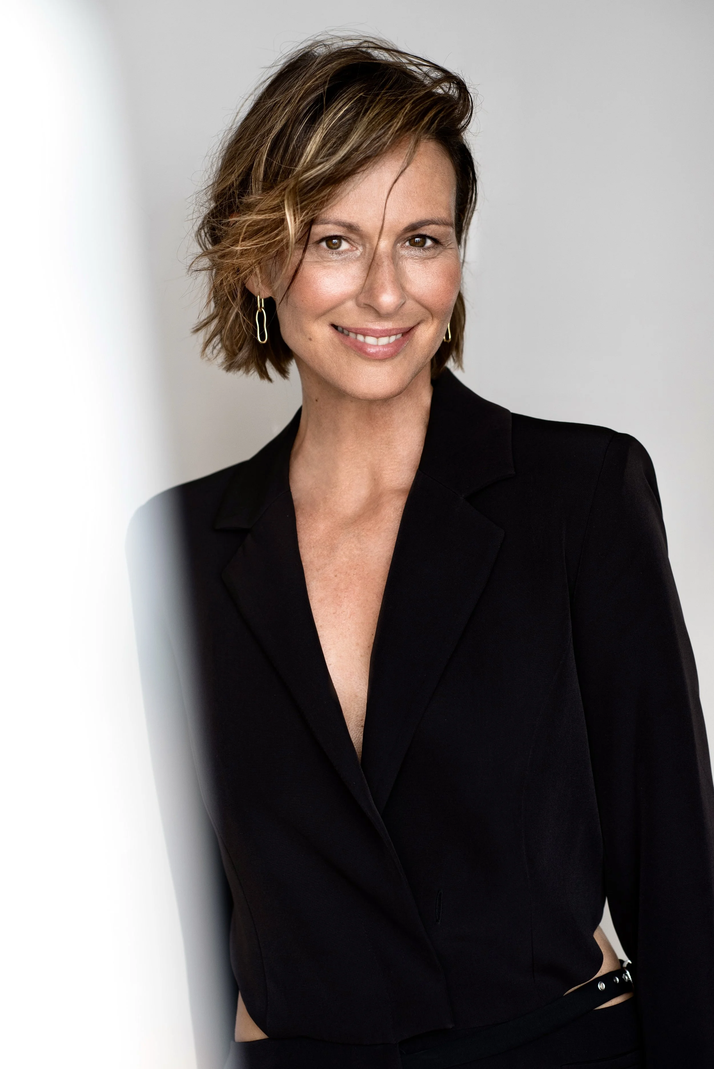 Porträt einer lächelnden Frau mit kurzem Haar, schwarzer Jacke und goldenen Ohrringen, stehend vor einem weißen Hintergrund.