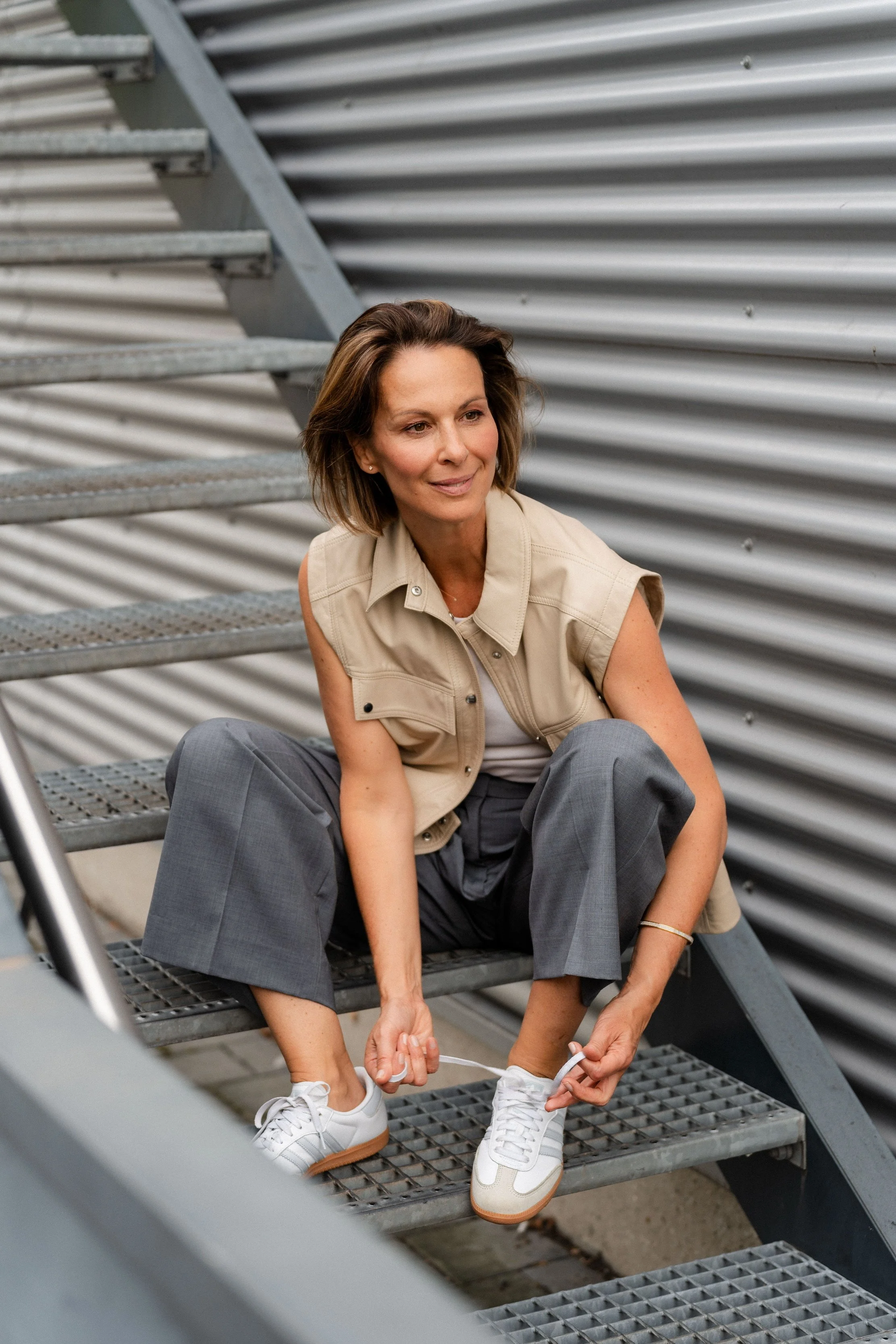 Frau auf Stufen, bindet Schuhe, trägt beige Weste und graue Hose, vor metallischer Wand.