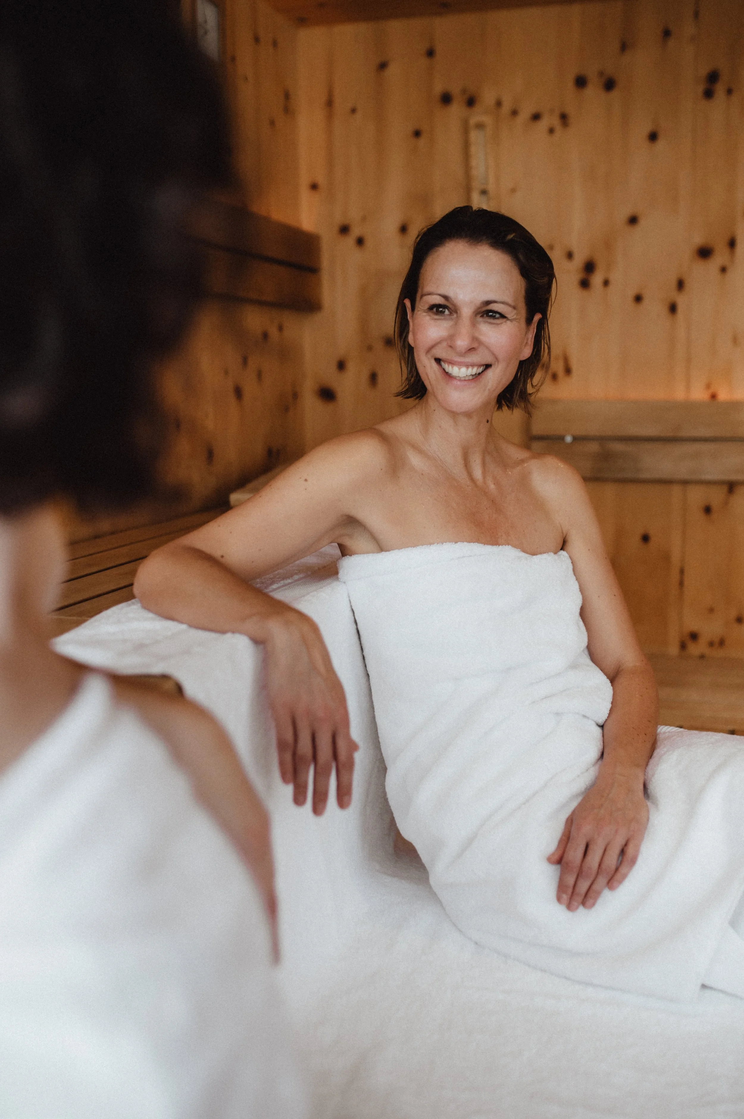 Zwei Frauen sitzen nackt und in weißen Handtüchern in einer Sauna, entspannen und lachen.