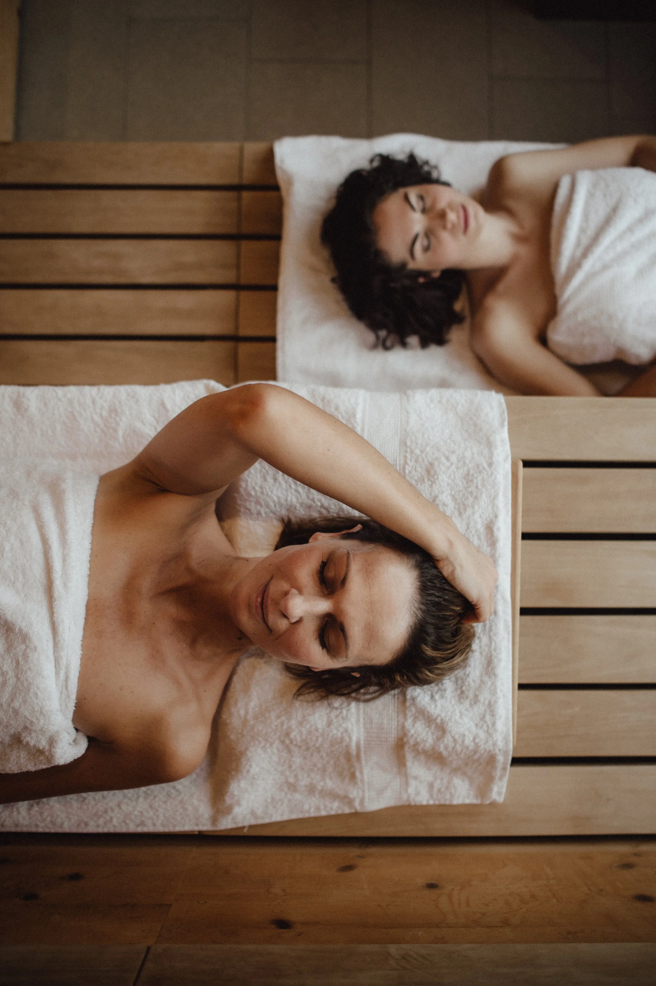 Zwei Frauen liegen entspannt in einem Spa oder Wellnessbereich, beide haben Handtücher um die Schultern gewickelt, die eine Frau mit gestreiftem Haar scheint die andere Frau in einem Moment der Ruhe zu sein.