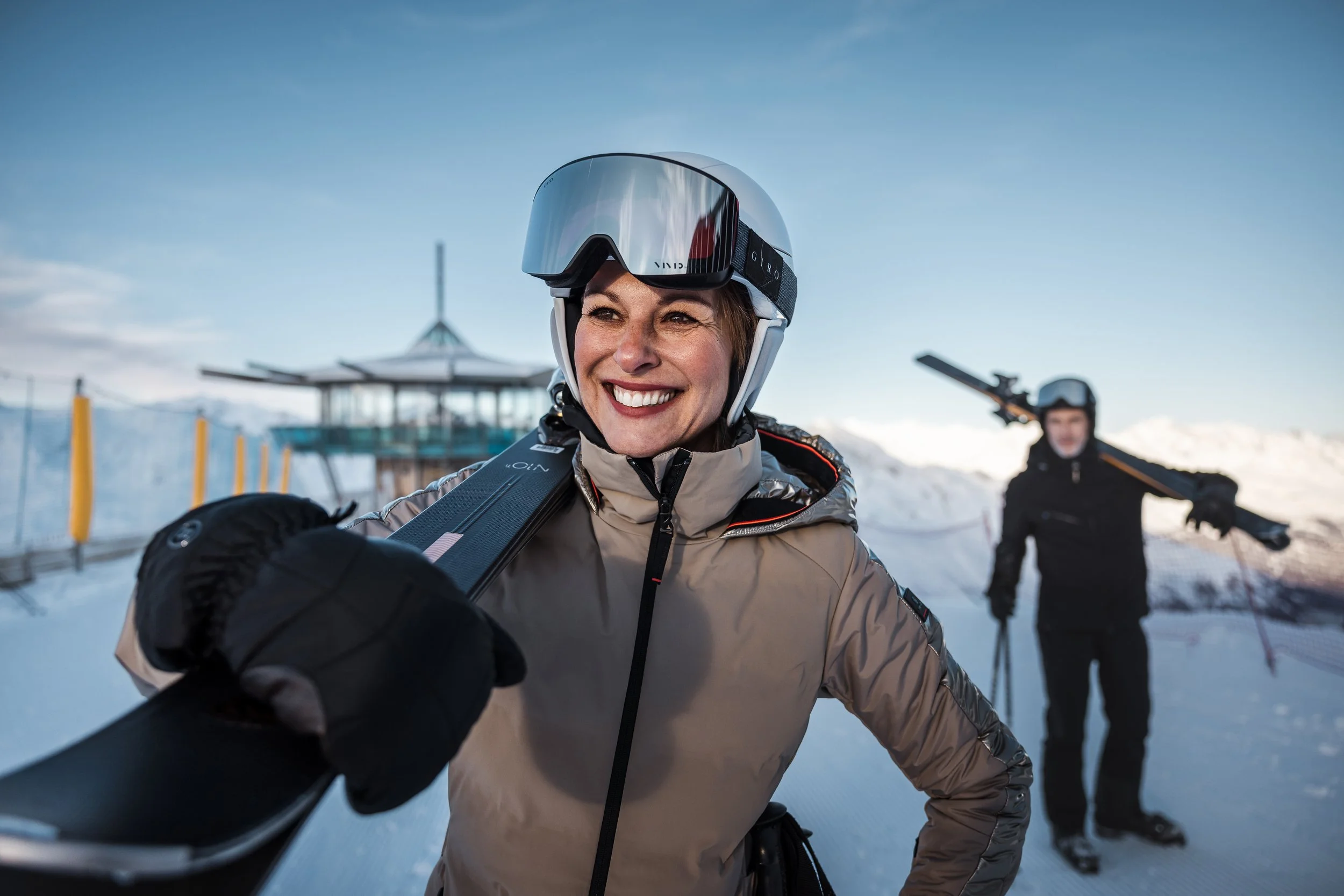 Eine smiling Frau in Skibekleidung nimmt ein Selfie mit Skiern auf einer Piste auf, im Hintergrund steht ein Mann mit Skischuhen, Skistöcken und Skiern, in einer winterlichen Berglandschaft bei Sonnenuntergang.
