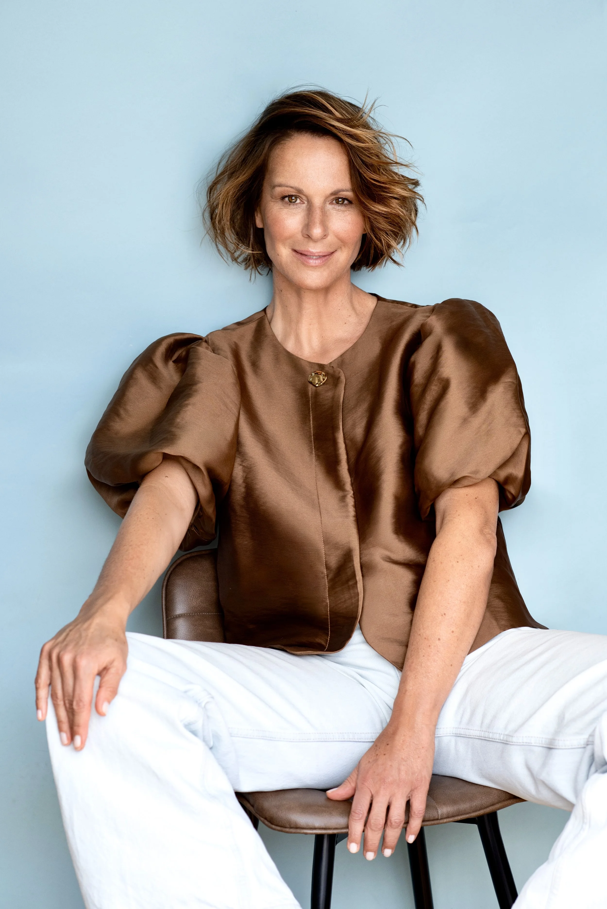 Eine Frau mit kurzen, lockigen braunen Haaren sitzt auf einem Stuhl vor blauer Wand, trägt eine braune Satin-Bluse mit Puffärmeln und weiße Hosen, und lächelt freundlich in die Kamera.