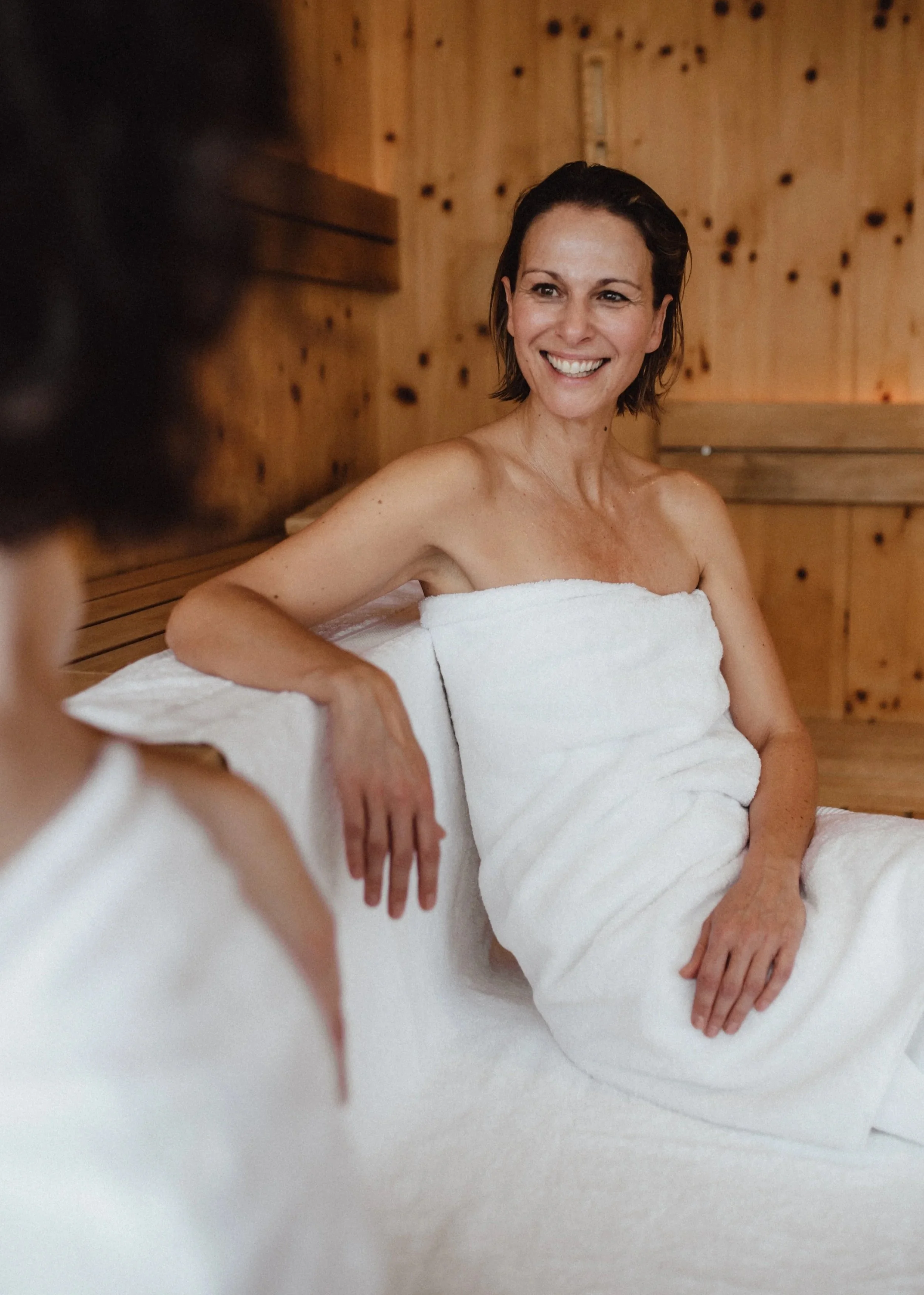 Zwei Frauen sitzen in einer Sauna, beide in Handtüchern gewickelt, lächeln und unterhalten sich.