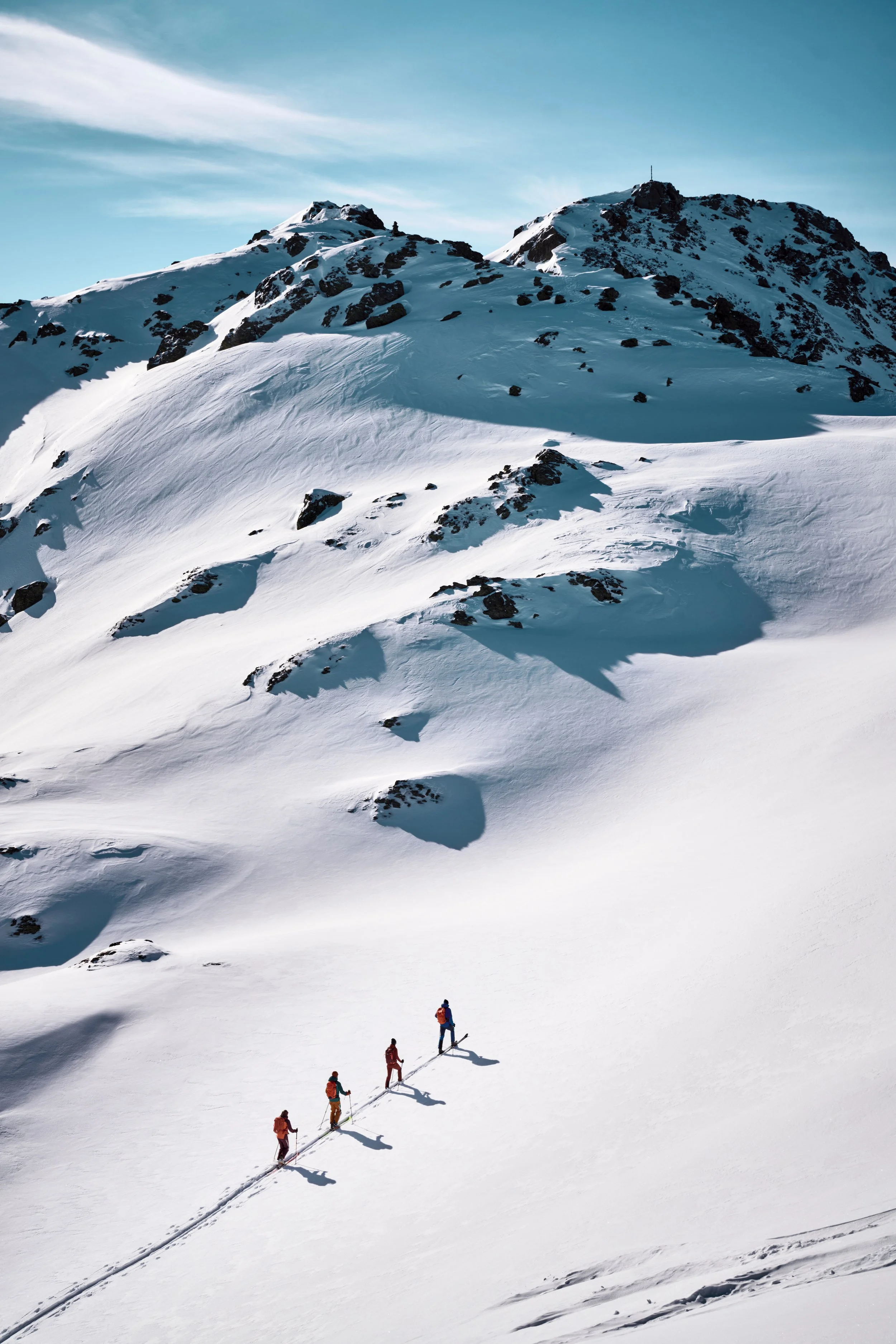 Vier Schneeschuhwanderer in bunter Kleidung wandern in einer verschneiten Berglandschaft bei sonnigem Himmel.