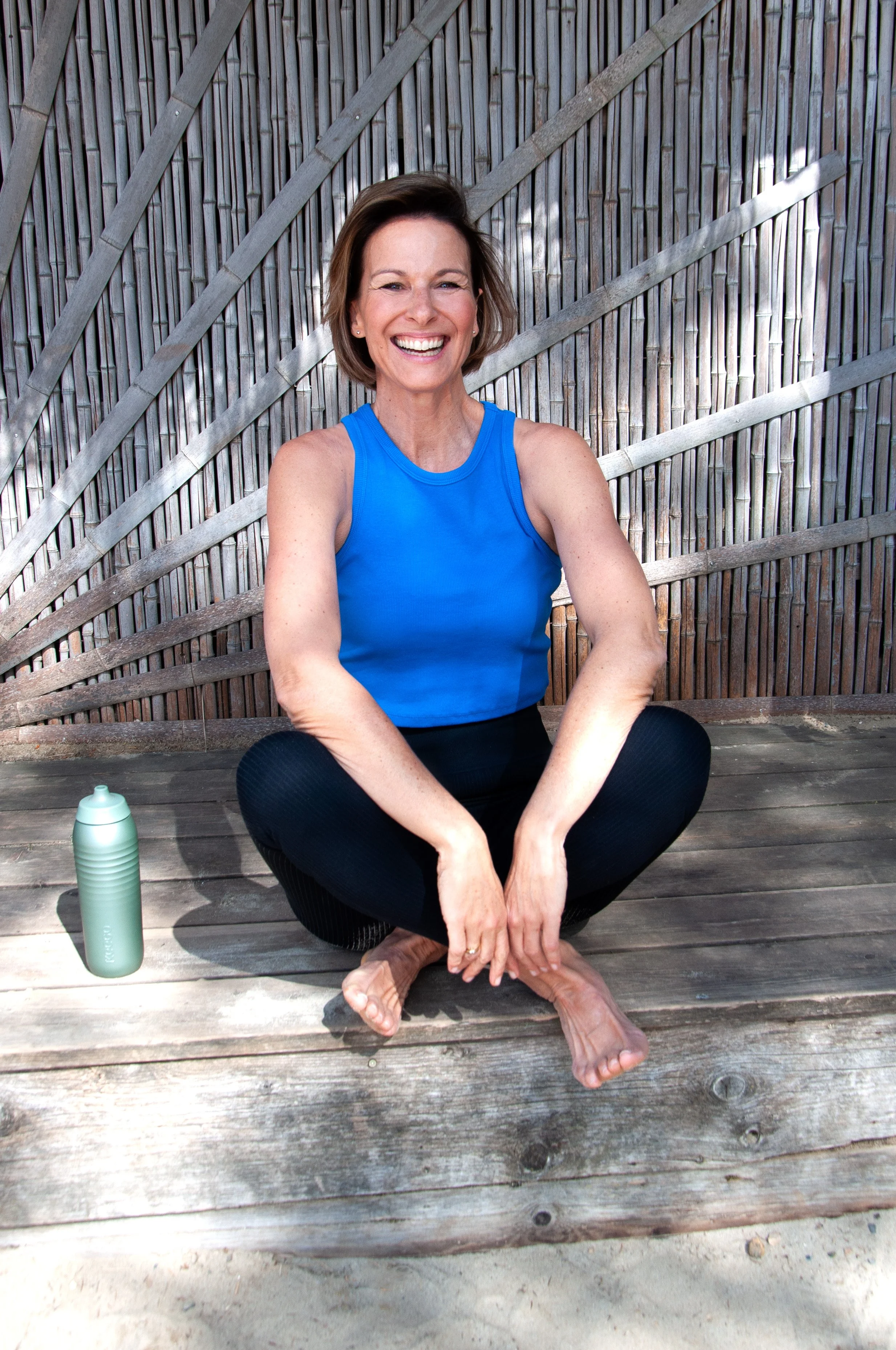 Freudige Frau im blauen Sporttop sitzt barfuß auf einer Holzbank, neben einer grünen Wasserflasche, vor einem Holzplatten-Hintergrund.