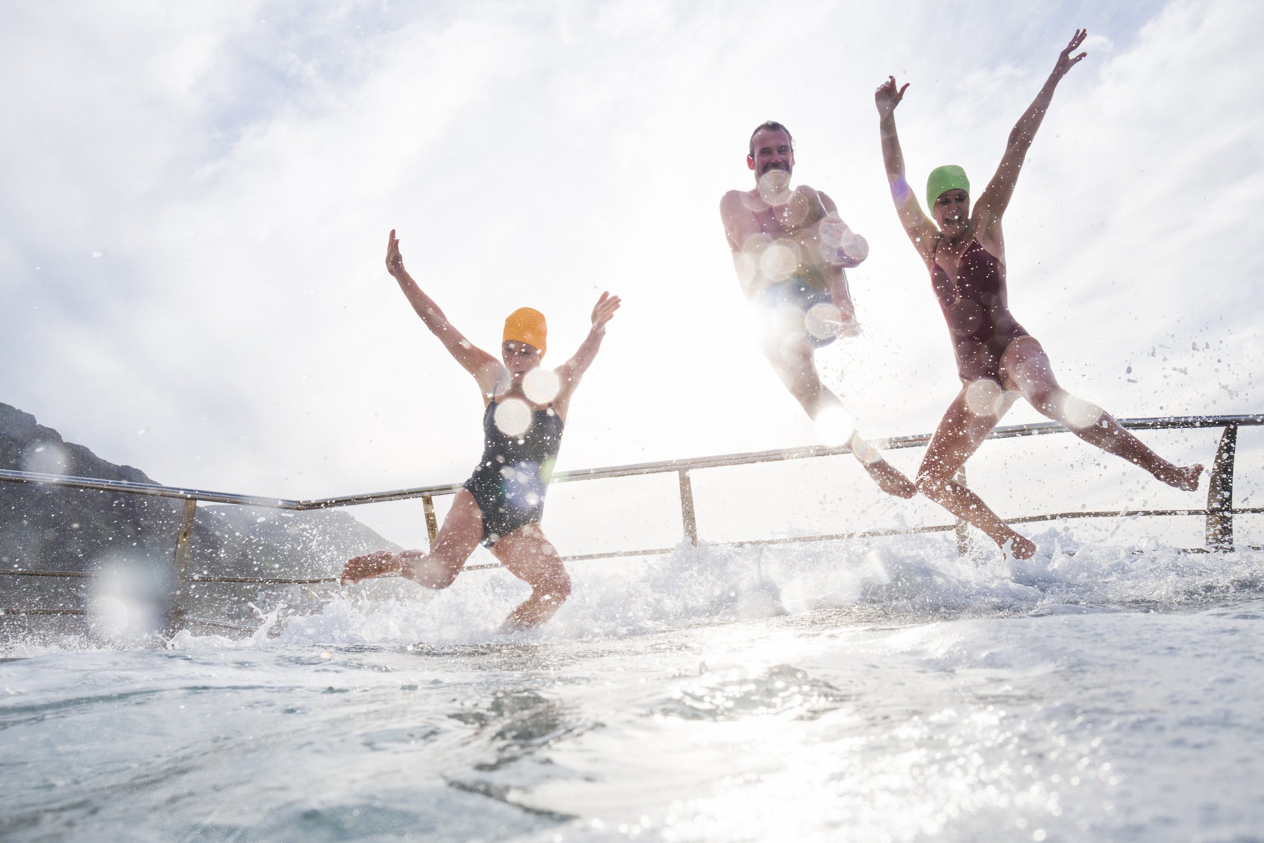 Drei Menschen in Badekleidung springen ins Wasser am Strand, lachen und haben Spaß bei sonnigem Wetter.