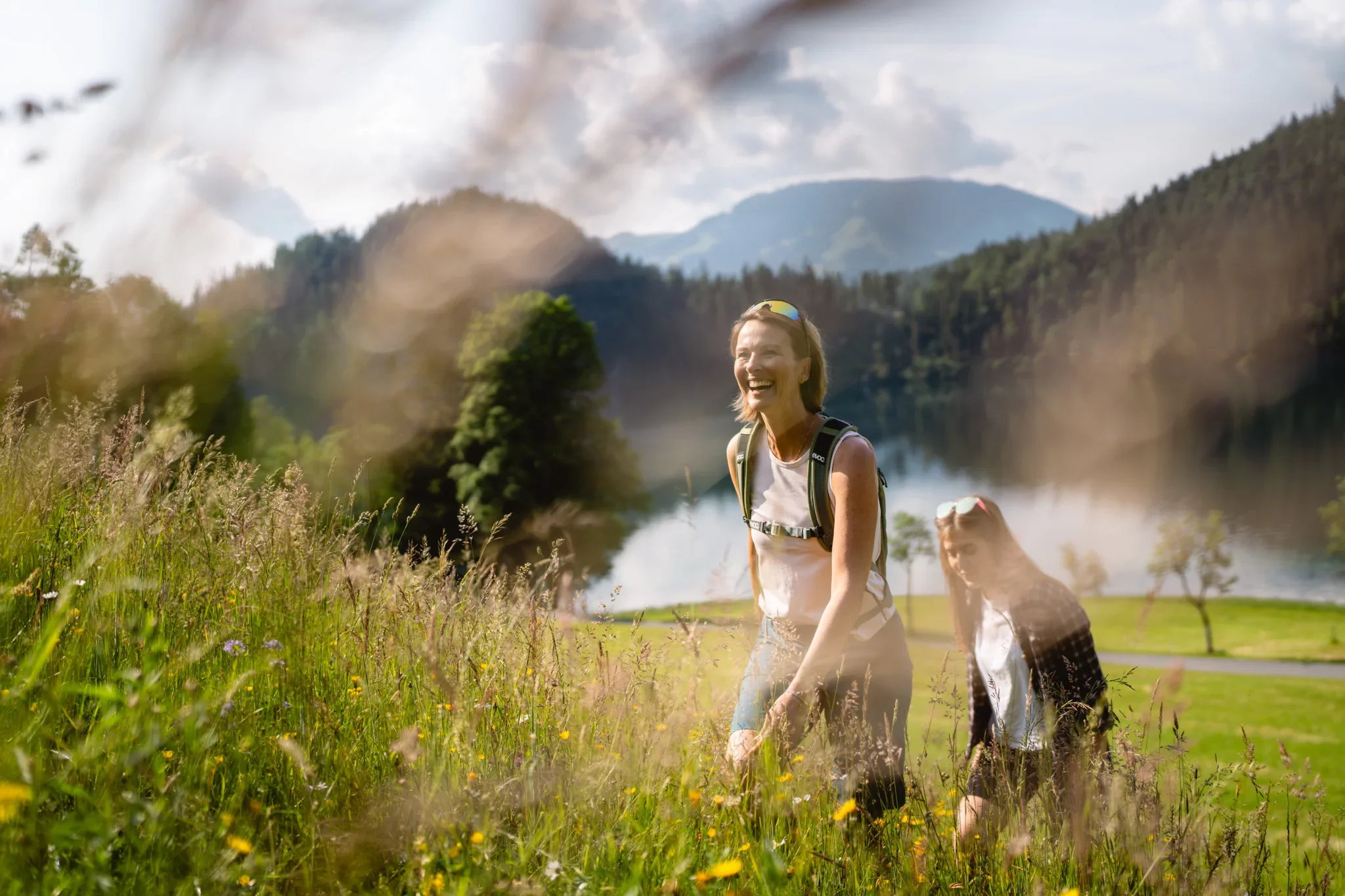 Zwei Frauen wandern an einem See in einer grünen Landschaft mit Bergen im Hintergrund bei sonnigem Wetter.