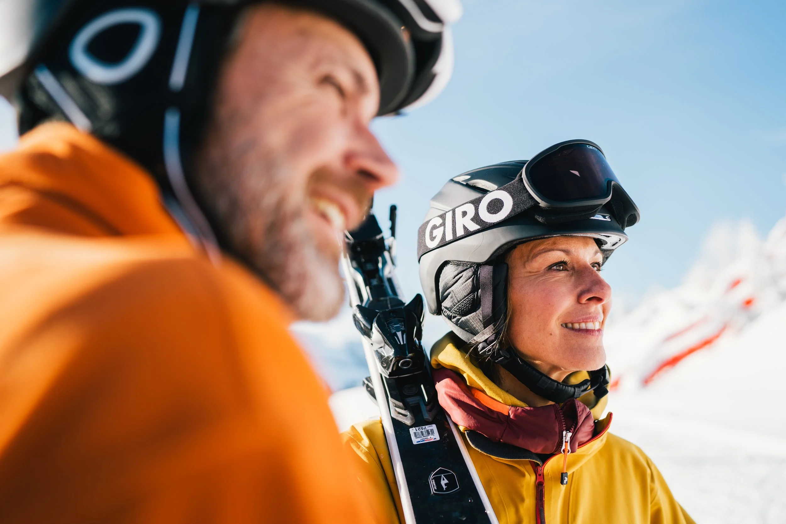 Zwei Skifahrer in Helm und Skibrille auf verschneitem Berg, lachend, blauer Himmel.