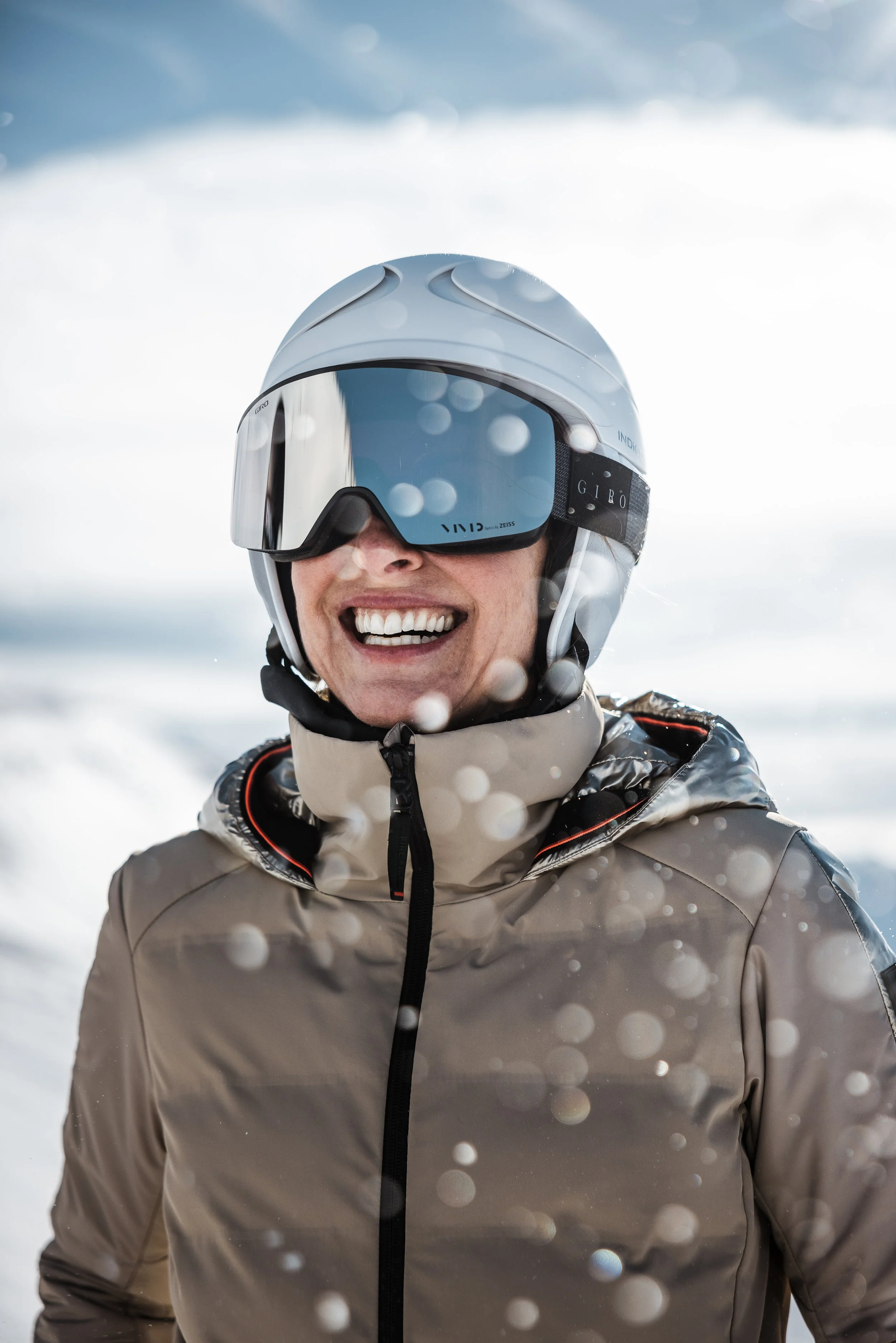 Person in Skijacke und Helm mit Skibrille, lachend im Schnee, im Wintersportgebiet.