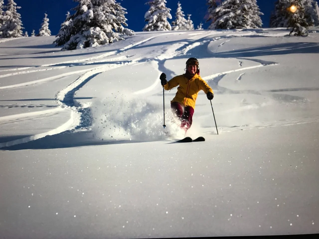 Person in gelber Jacke und schwarzen Helm beim Skifahren im tiefen Schnee in den Bergen, sonniger Himmel, verschneite Bäume im Hintergrund.