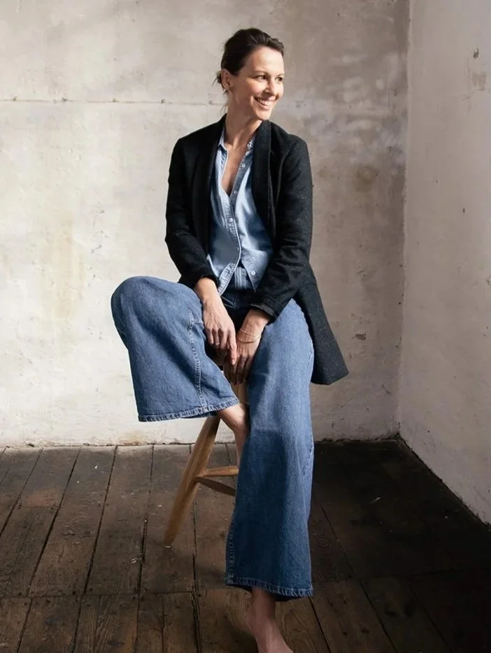 Eine lachende Frau sitzt auf einem Hocker vor einer blanken, hellen Wand, trägt eine dunkle Jacke, ein blauer Hemd und weite Jeans, barfuß, in einem natürlichen, minimalistischen Raum.