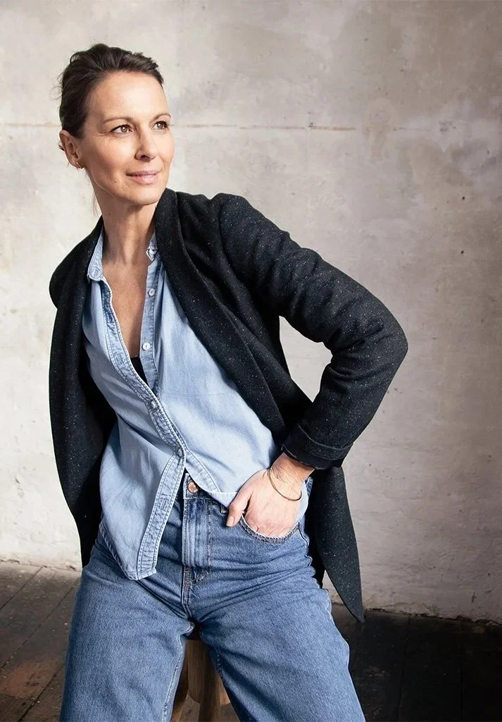 Eine Frau sitzt vor einer hellen, unebenen Wand und schaut nach rechts. Sie trägt eine dunkelglänzende Jacke, eine hellblaue Jeansbluse und Jeans. Sie hat kurze, dunkle Haare und sieht entspannt und selbstbewusst aus.