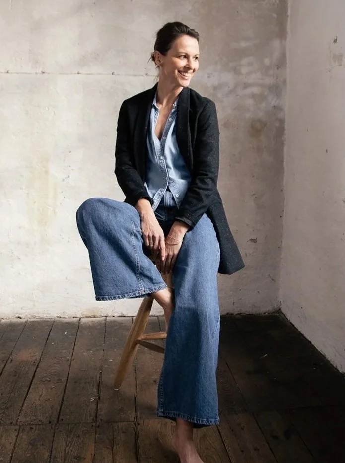 Eine Frau sitzt barfuß auf einem Holzstuhl vor einer weißen Wand, trägt Jeans, ein blaugraues Hemd und eine schwarze Jacke, lächelt.