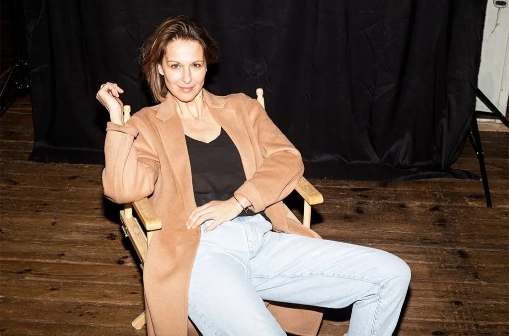 Eine Frau sitzt entspannt in einem Holzsessel vor einem schwarzen Hintergrund, trägt eine braune Jacke, ein schwarzes Oberteil und helle Jeans.