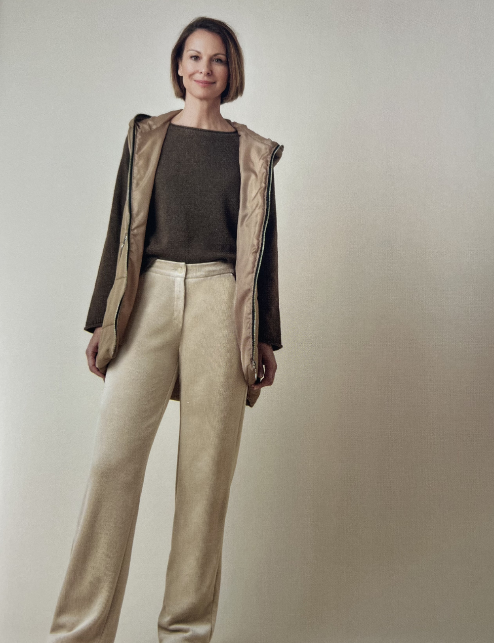 Eine Frau trägt einen braunen Pullover, eine beige Weste mit Kapuze, und beige weite Hosen.