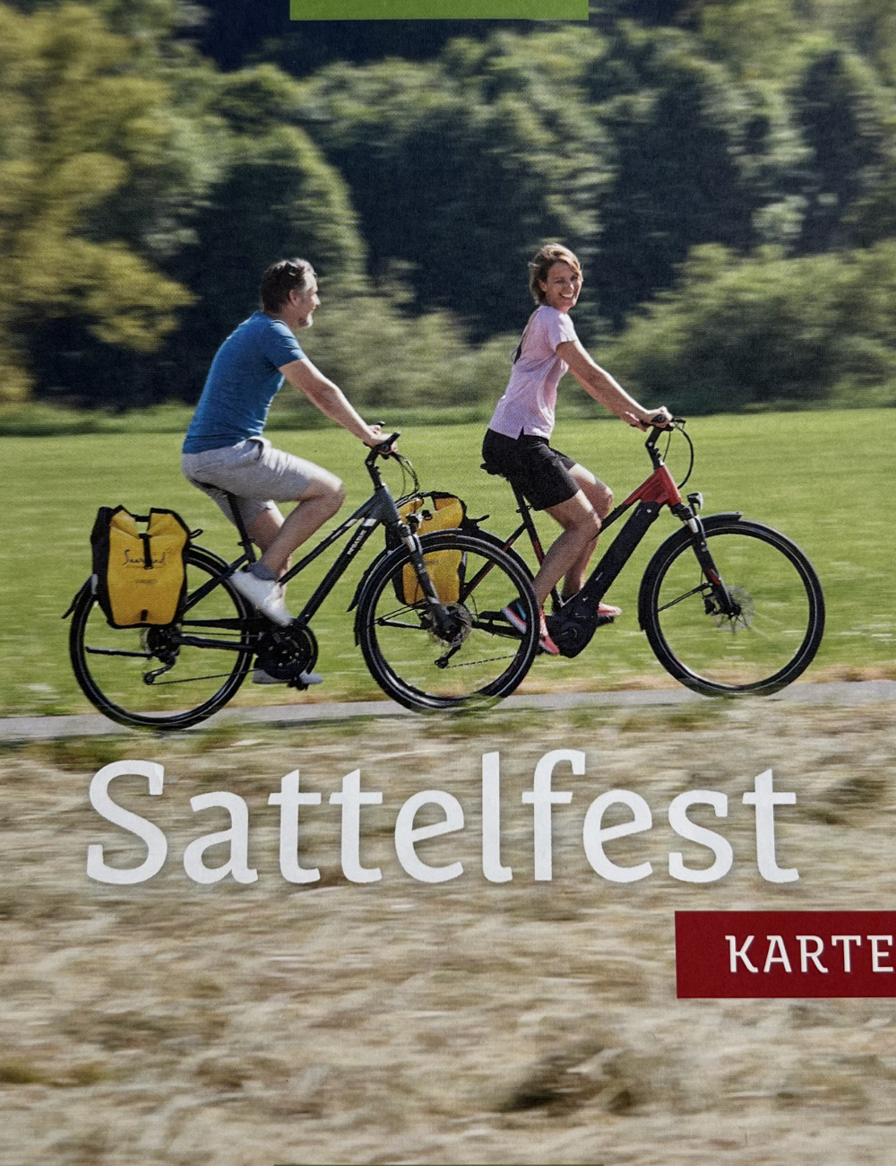 Zwei Frauen fahren Fahrrad auf einem Feld mit Bäumen im Hintergrund, lachen und haben Spaß bei Sattelfest.