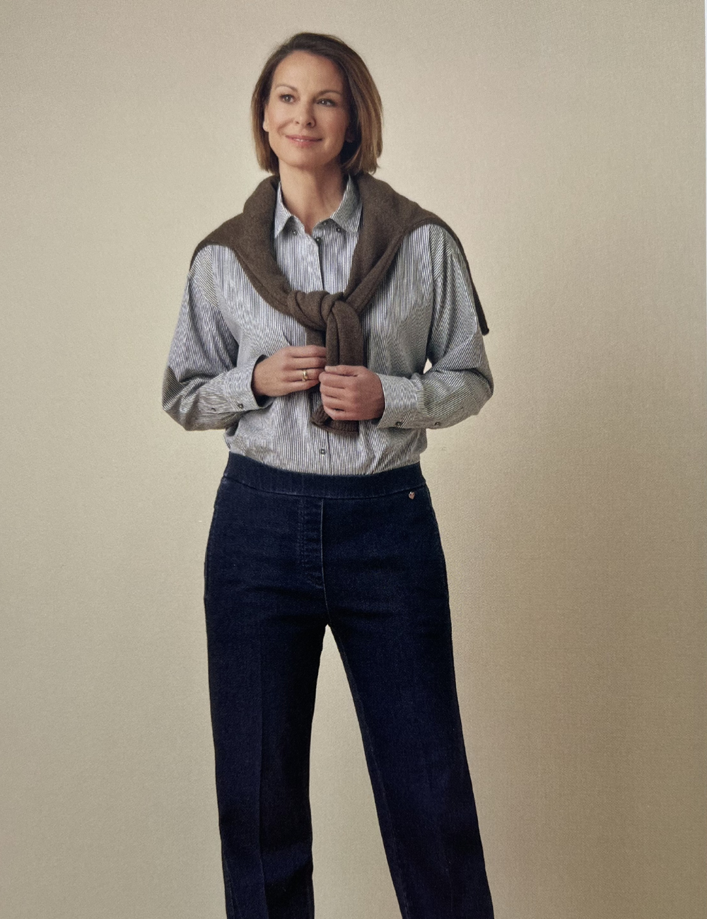 Frau in Hemd mit Pullover über den Schultern vor hellem Hintergrund.