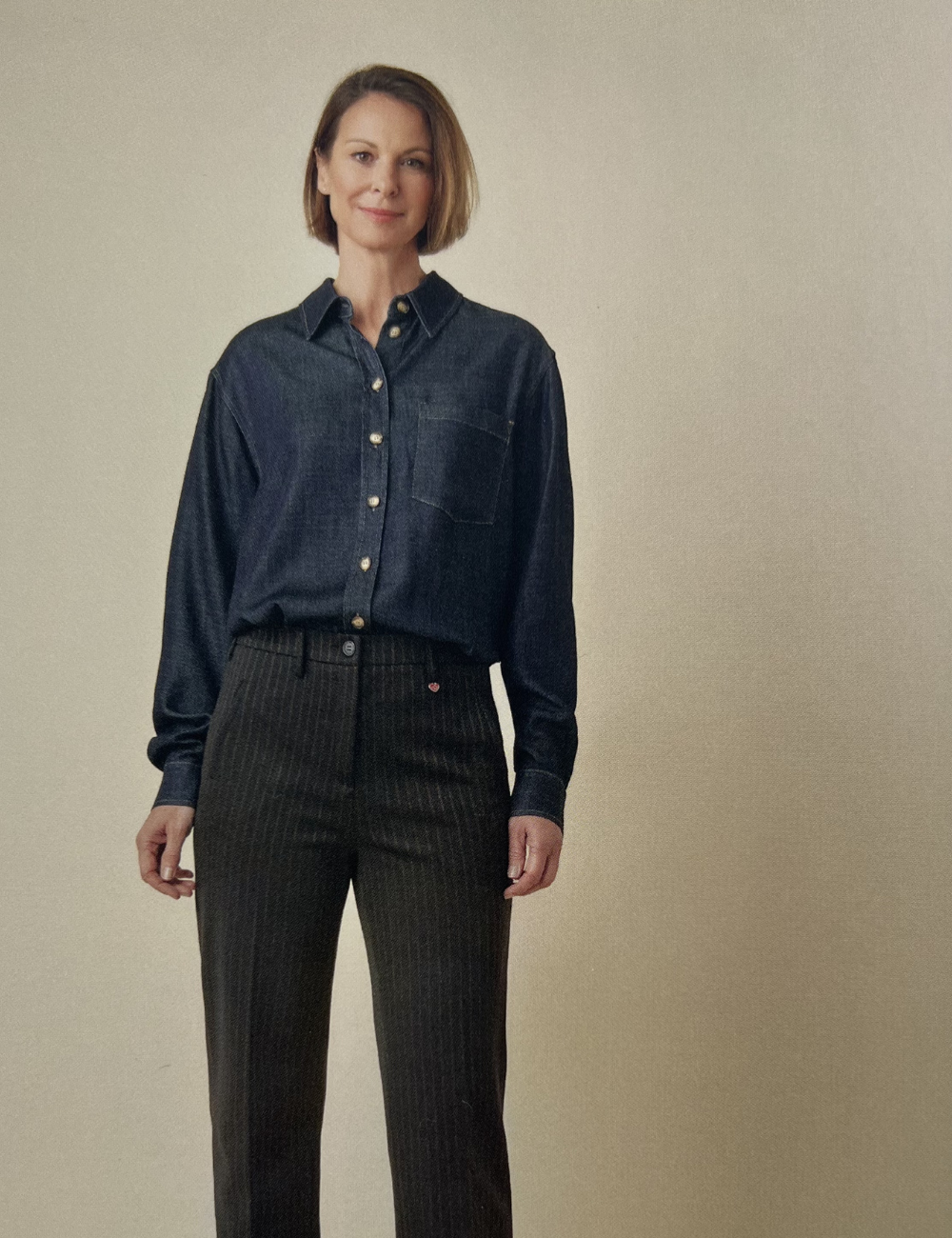 Eine Frau trägt eine dunkelblaue Jeansbluse und schwarze Hose mit feinem Streifenmuster, vor einem beigen Hintergrund.
