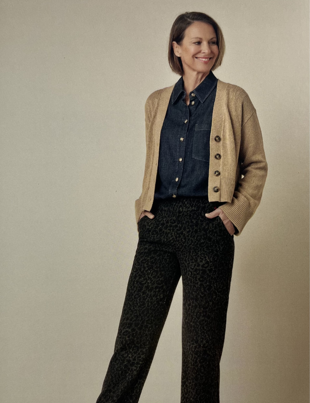 Frau im Business casual Outfit, trägt eine dunkelblaue Bluse, eine beige Strickjacke und schwarze Hosen mit Leopardenmuster, lächelnd und vor neutralem Hintergrund stehend.