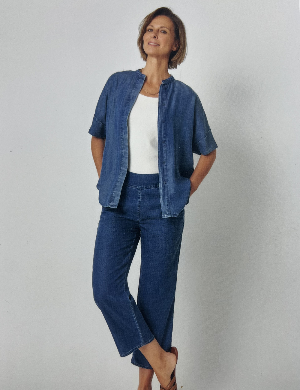 Eine Frau in einer Jeans-Jeansjacke und weißem T-Shirt steht vor einer weißen Wand und lächelt.