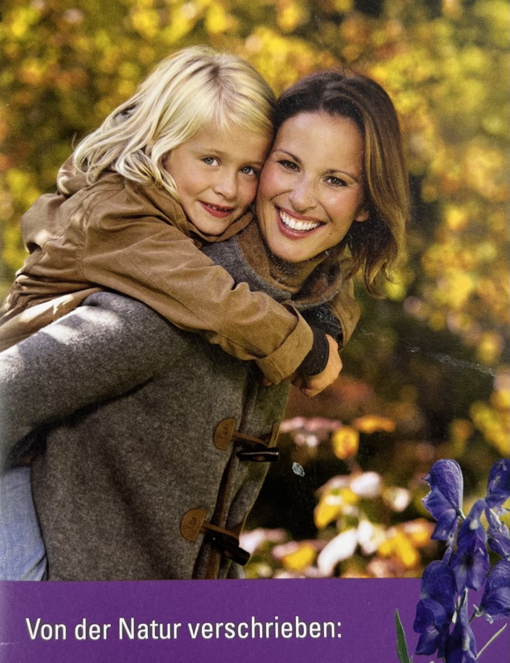 Eine Frau trägt ein lachendes Mädchen auf dem Rücken, beide umarmt, im Park mit herbstlichen Bäumen, im Vordergrund Blumen. Text: "Von der Natur verschrieben".