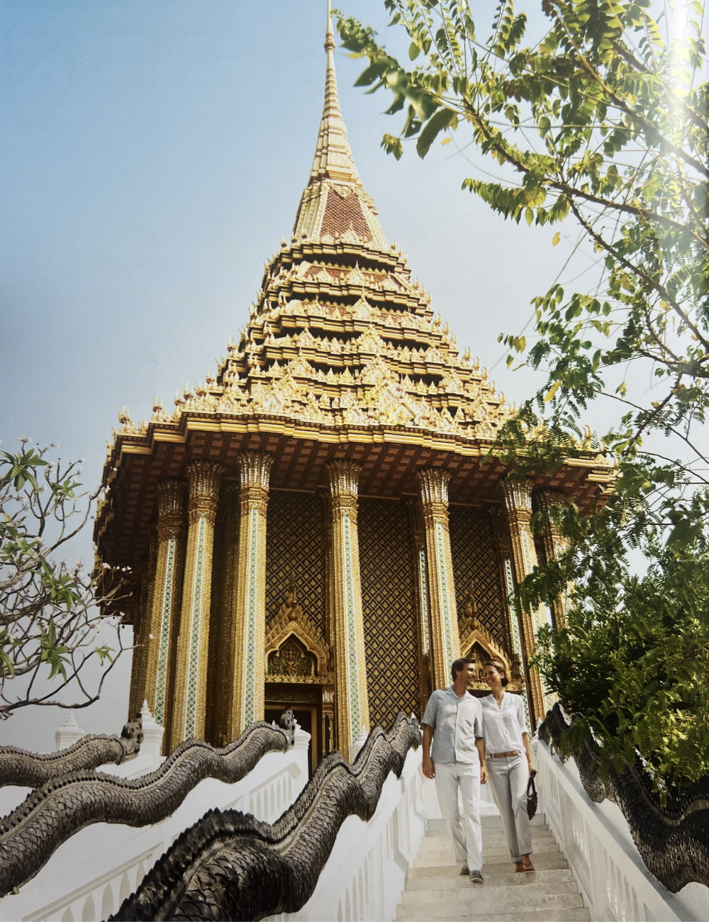 Ein goldener Tempel in Thailand mit zwei Menschen, die die Treppe hinuntergehen, umgeben von grünen Bäumen bei sonnigem Wetter.
