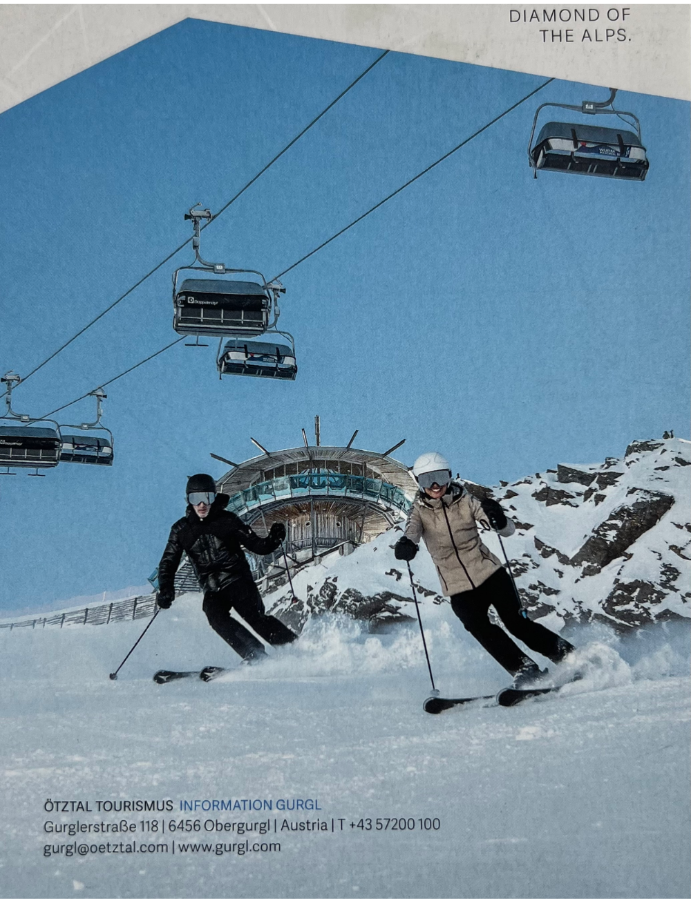 Zwei Skifahrer auf schneebedecktem Berg mit einer modernen Berghütte im Hintergrund, Seilbahn mit Gondeln oberhalb, klare blauer Himmel, Wintersport in den Alpen, Kontaktinformationen für Ötztal Tourismus