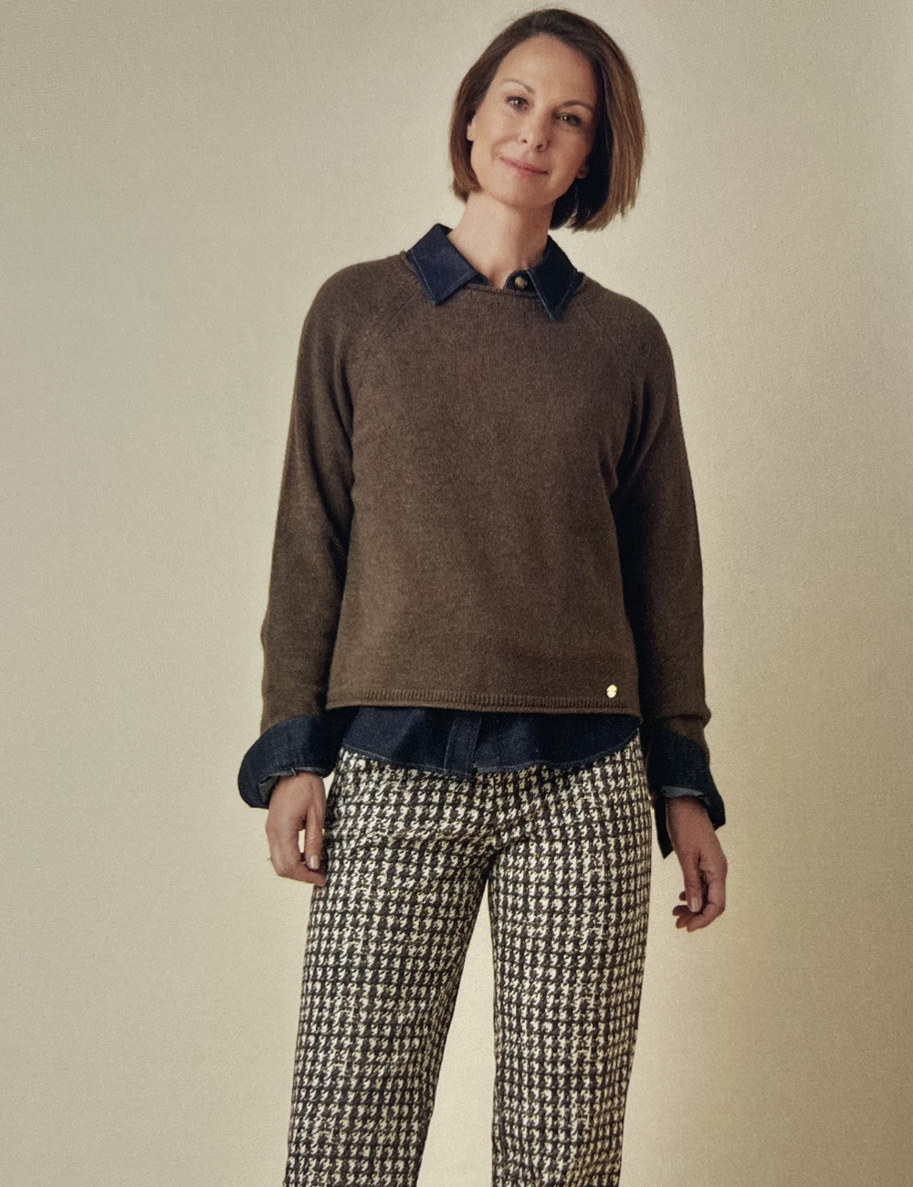 Frau mit braunem Pullover, rotem Haarguide und karierten Hosen, vor einem neutralen Hintergrund.