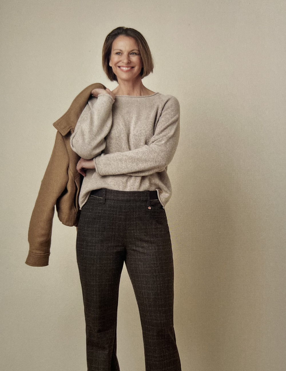 Eine lachende Frau mit kurzen braunen Haaren, trägt einen beigen Pulli und dunkle, kariert aussehende Hosen, hält eine braune Jacke über die Schulter.