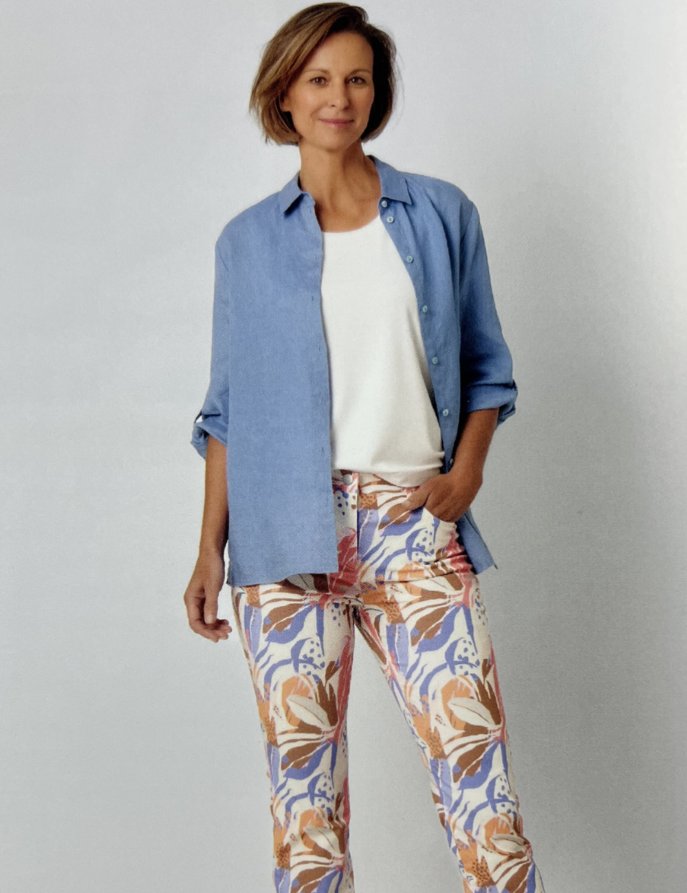 Eine Frau steht vor einem weißen Hintergrund, trägt ein weißes T-Shirt, eine offene blaue Hemdbluse und bunte, gemusterte Hosen mit Orange, Blau und Beige Tönen.