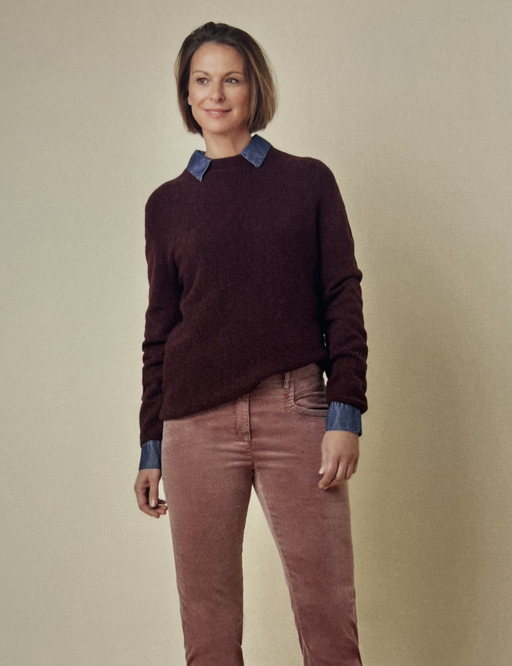 Eine Frau steht vor einer beige Wand, trägt eine dunkelbraune Pullover über einem blauen Hemd und rosa Hosen.