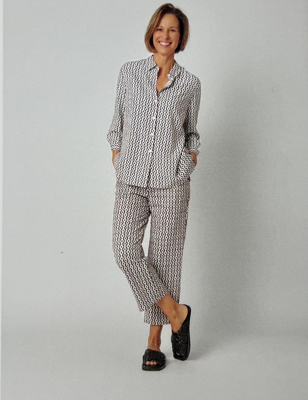 Frau in gemütlicher, gemusterter Hose und Hemd, lächelnd, schwarze Schuhe, steht vor weißem Hintergrund.