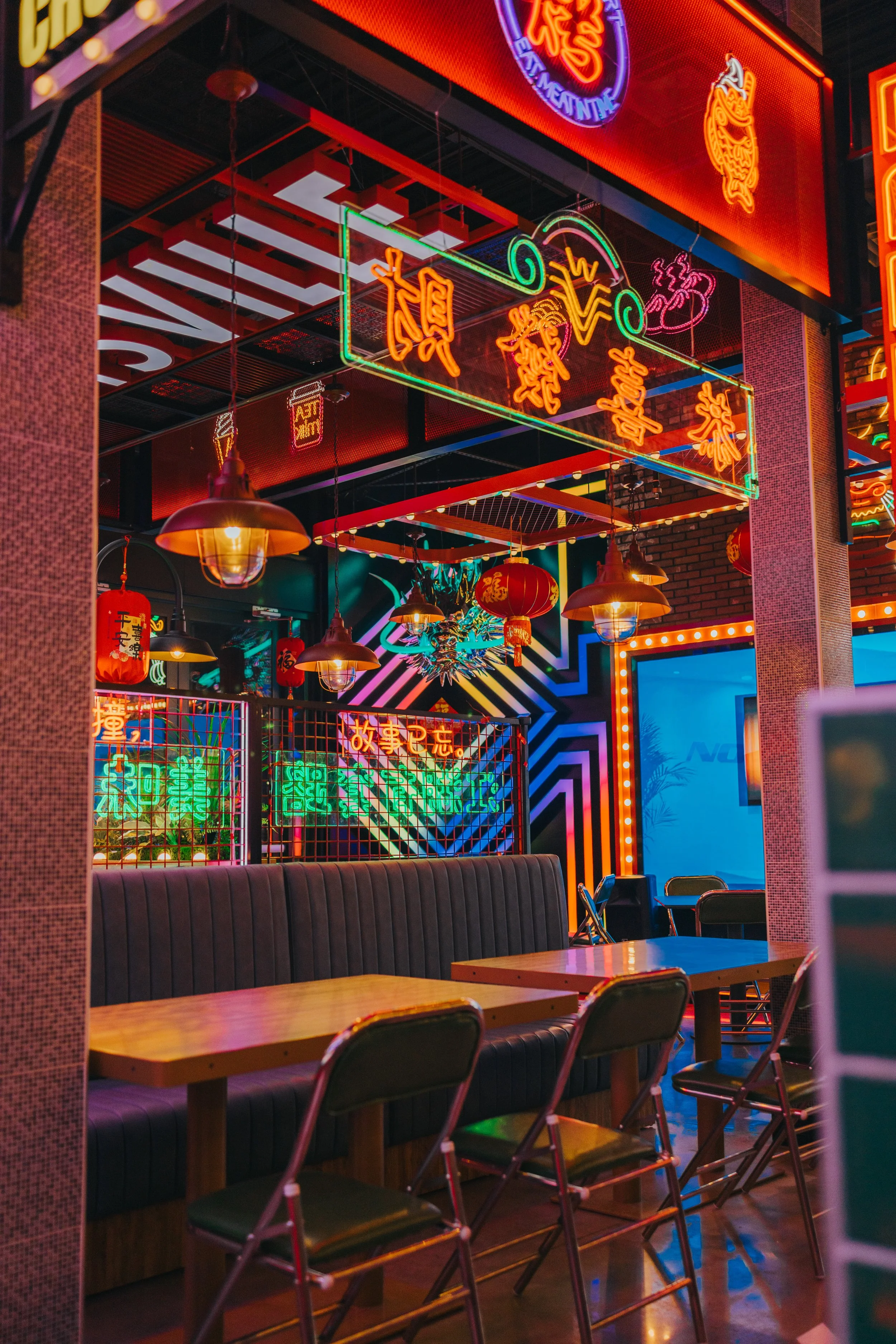 Interior of a restaurant or bar decorated with colorful neon signs and hanging red lanterns, featuring seating with wooden tables and black padded booths.