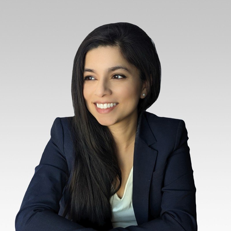 A young woman with long black hair, wearing a navy blue blazer and a white top, smiling and looking to the side against a light gray background.