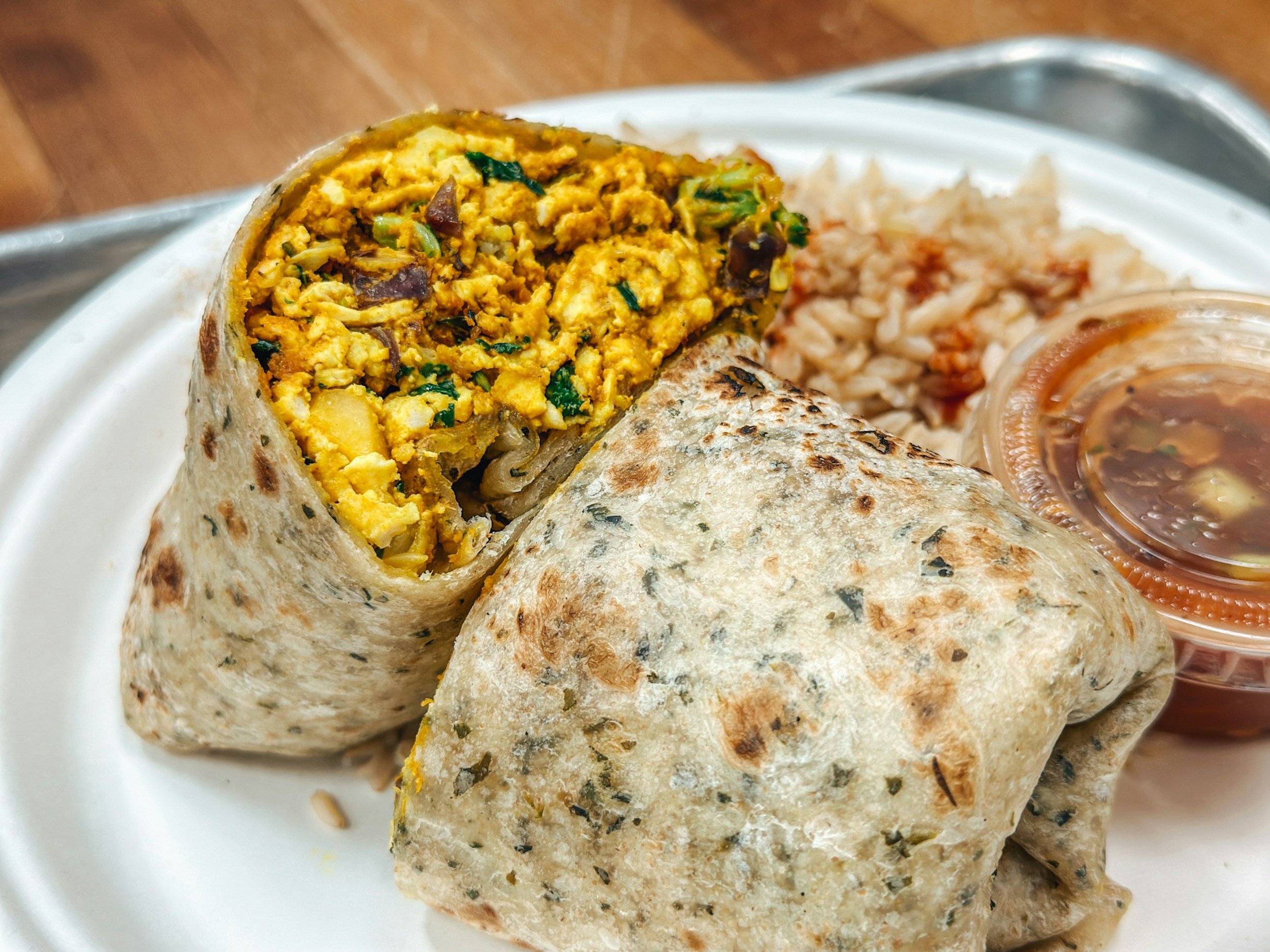 Close-up of a burrito filled with scrambled eggs, vegetables, and herbs, served with rice, salsa, and beans on a white plate.