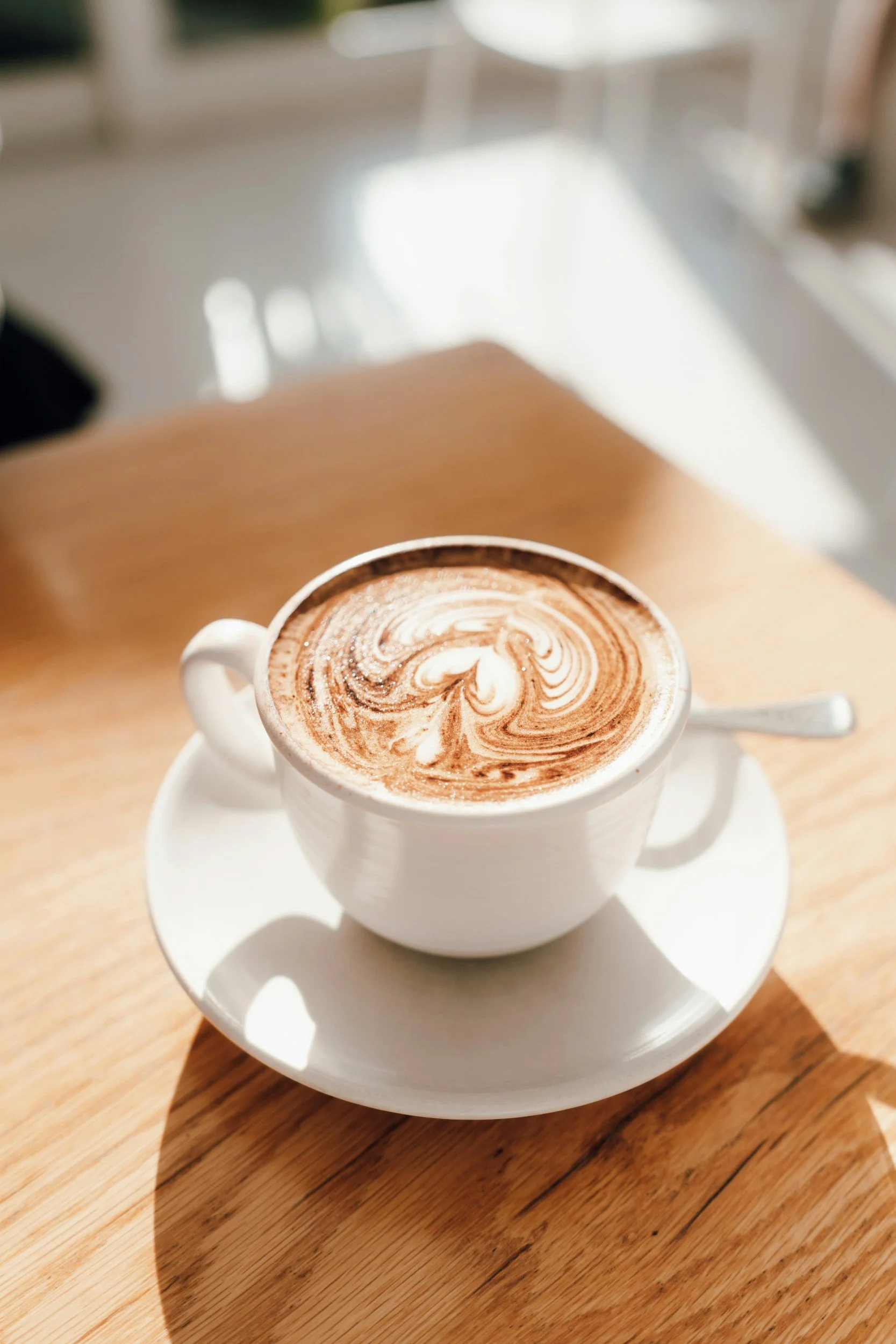 A white ceramic cup filled with a latte with swirled foam art, placed on a matching white saucer on a wooden table.