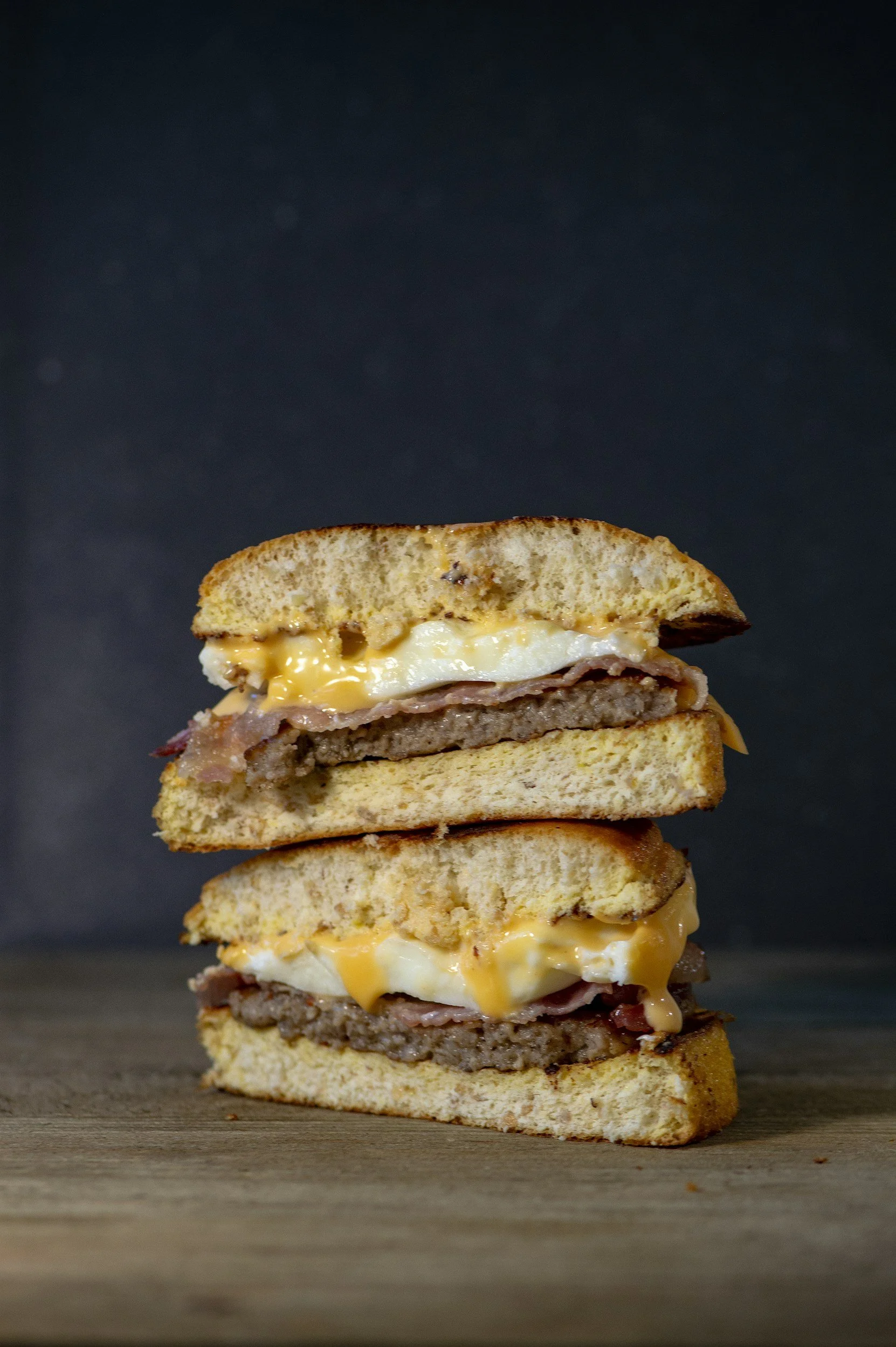 A double cheeseburger with two beef patties, bacon, fried egg, and melted cheese on toasted bread slices, placed on a wooden surface against a dark background.