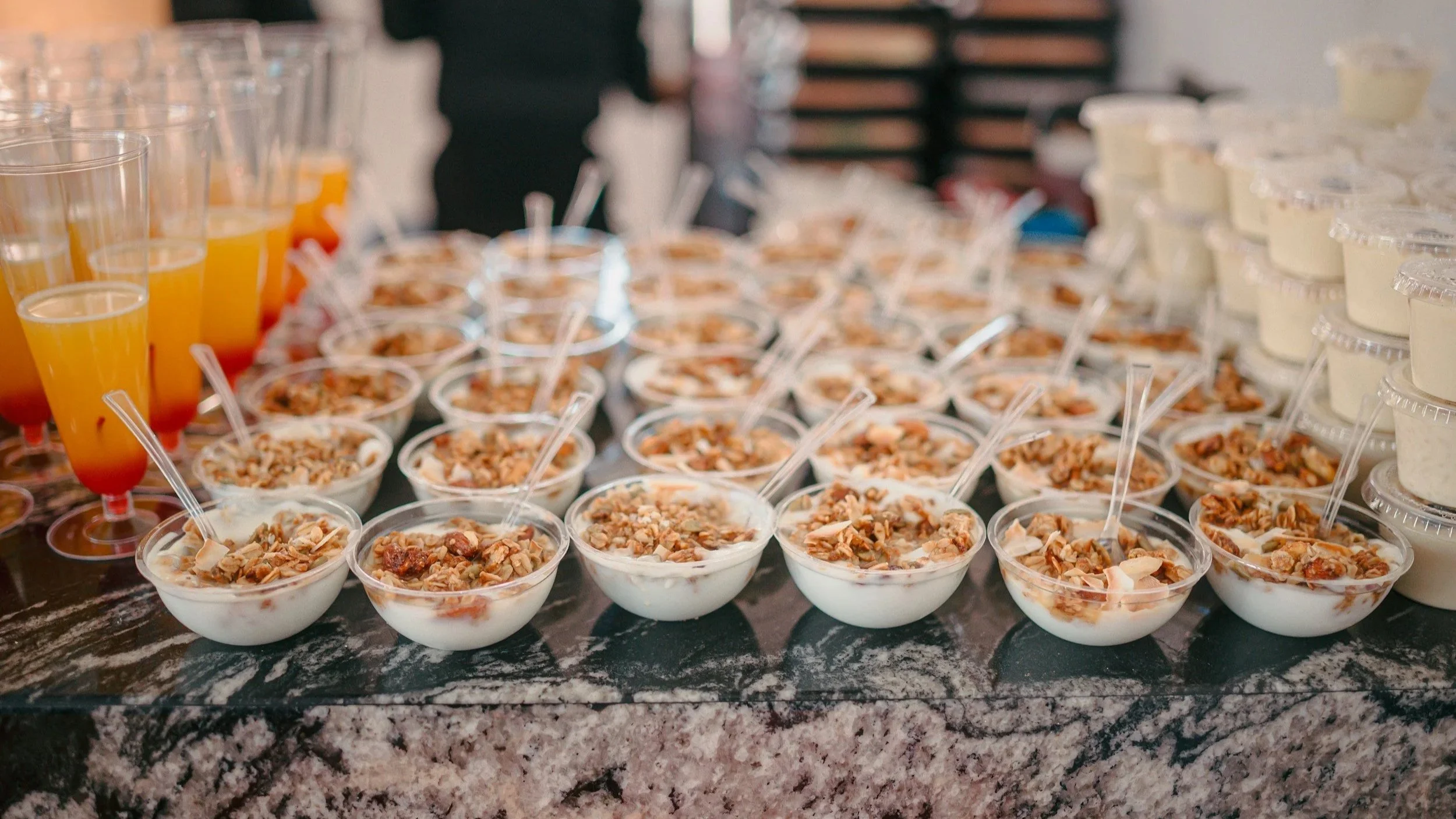 Various breakfast items on a buffet table, including small bowls of yogurt topped with nuts, cups of orange juice, and containers of other breakfast foods.