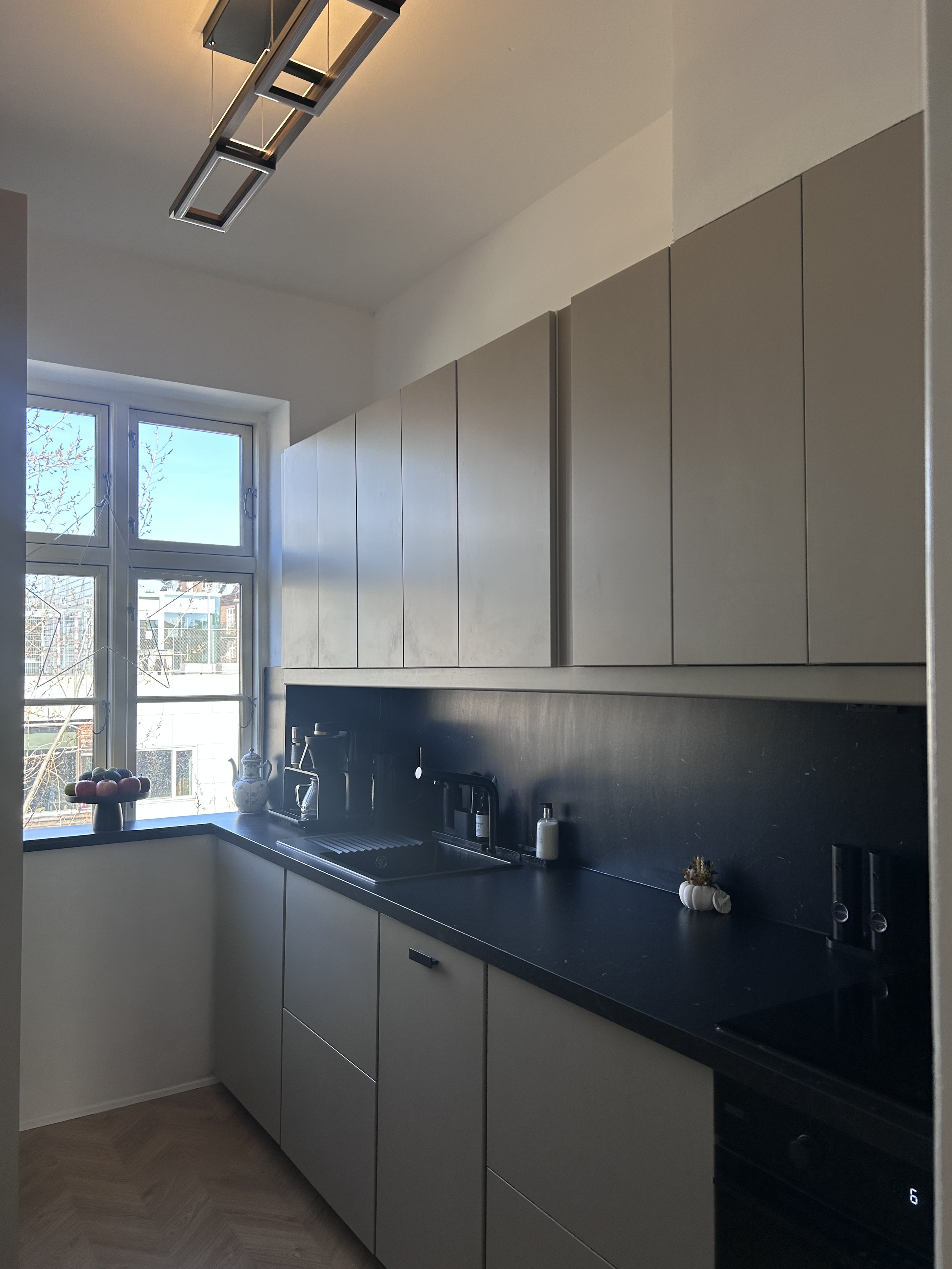 Modern kitchen with beige cabinets, black countertop, and a large window with a view of trees and neighboring buildings. Decor includes a bowl of apples, a teapot, a coffee maker, a soap dispenser, and small pumpkins on the counter.